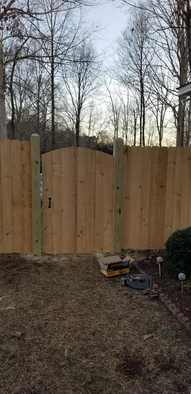 Wooden privacy fence with a curved gate in a leaf-covered backyard, bordered by bare trees.
