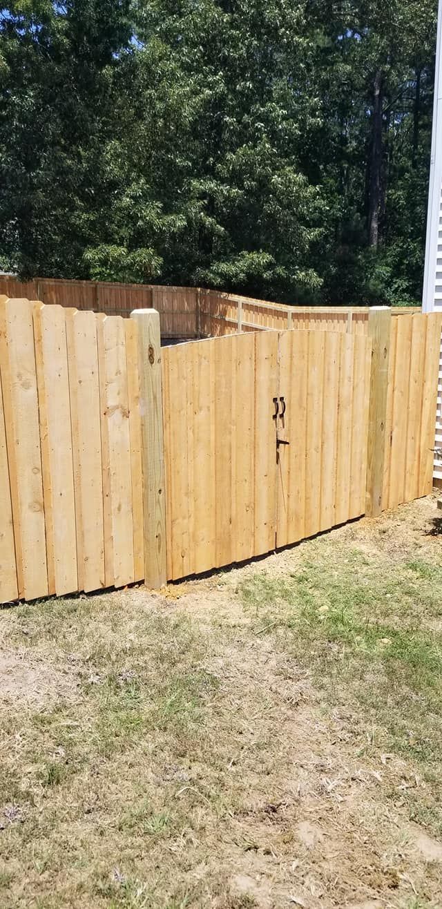Wooden backyard fence with a curved gate, bordered by dry grass and trees.