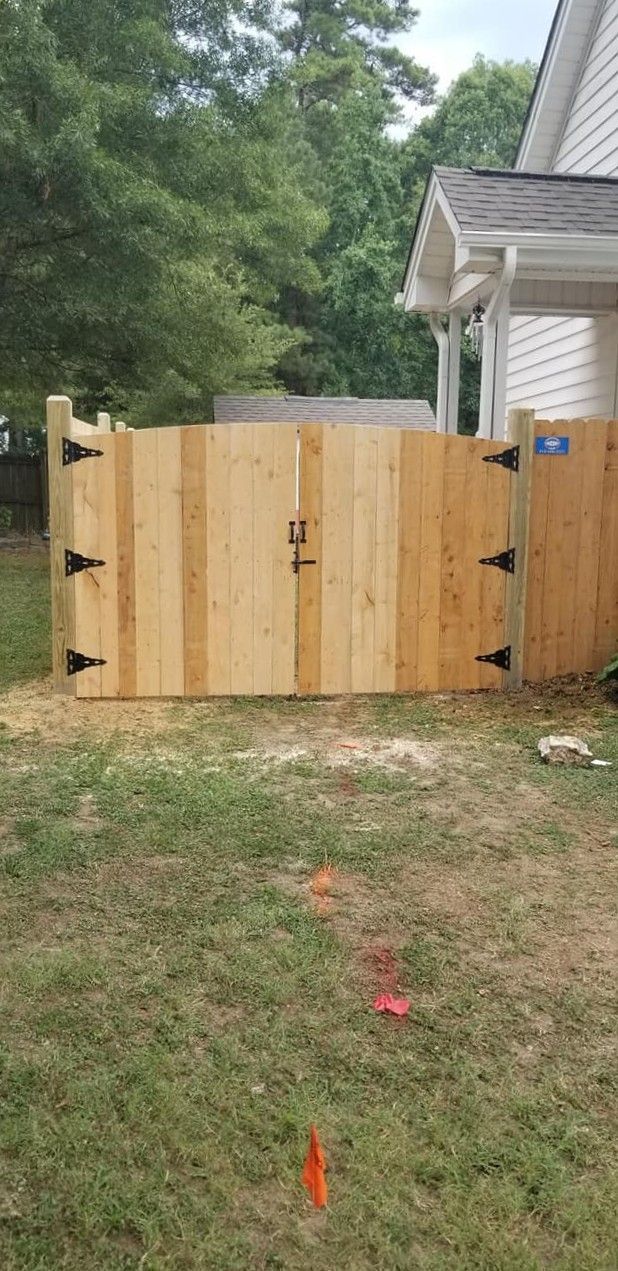 Wooden backyard gate with black hinges and latch, beside a white house and trees.