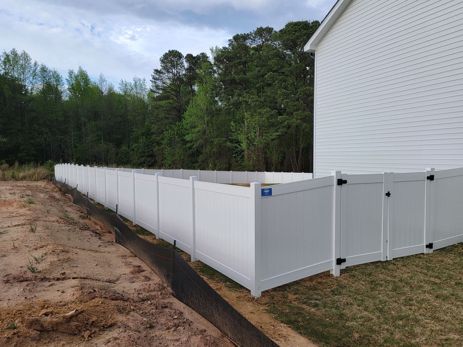 White vinyl privacy fence along a house beside a dirt lot and trees under a cloudy sky