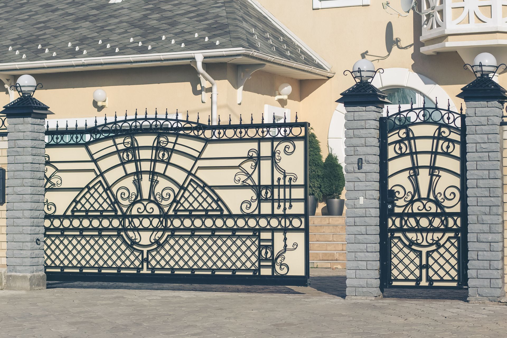 Ornate black wrought-iron gate and pedestrian door in front of a stone house with a tiled roof.
