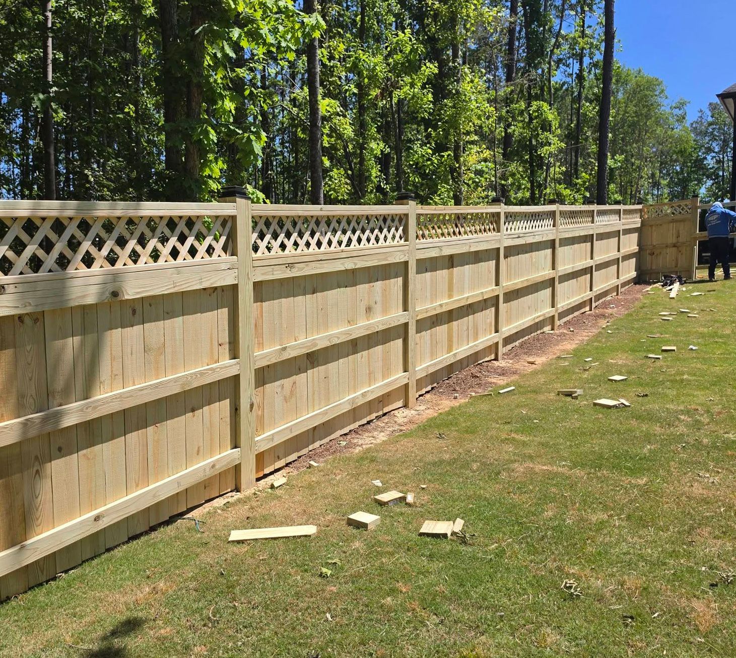 Long wooden privacy fence along a sunny grassy yard with trees in the background.