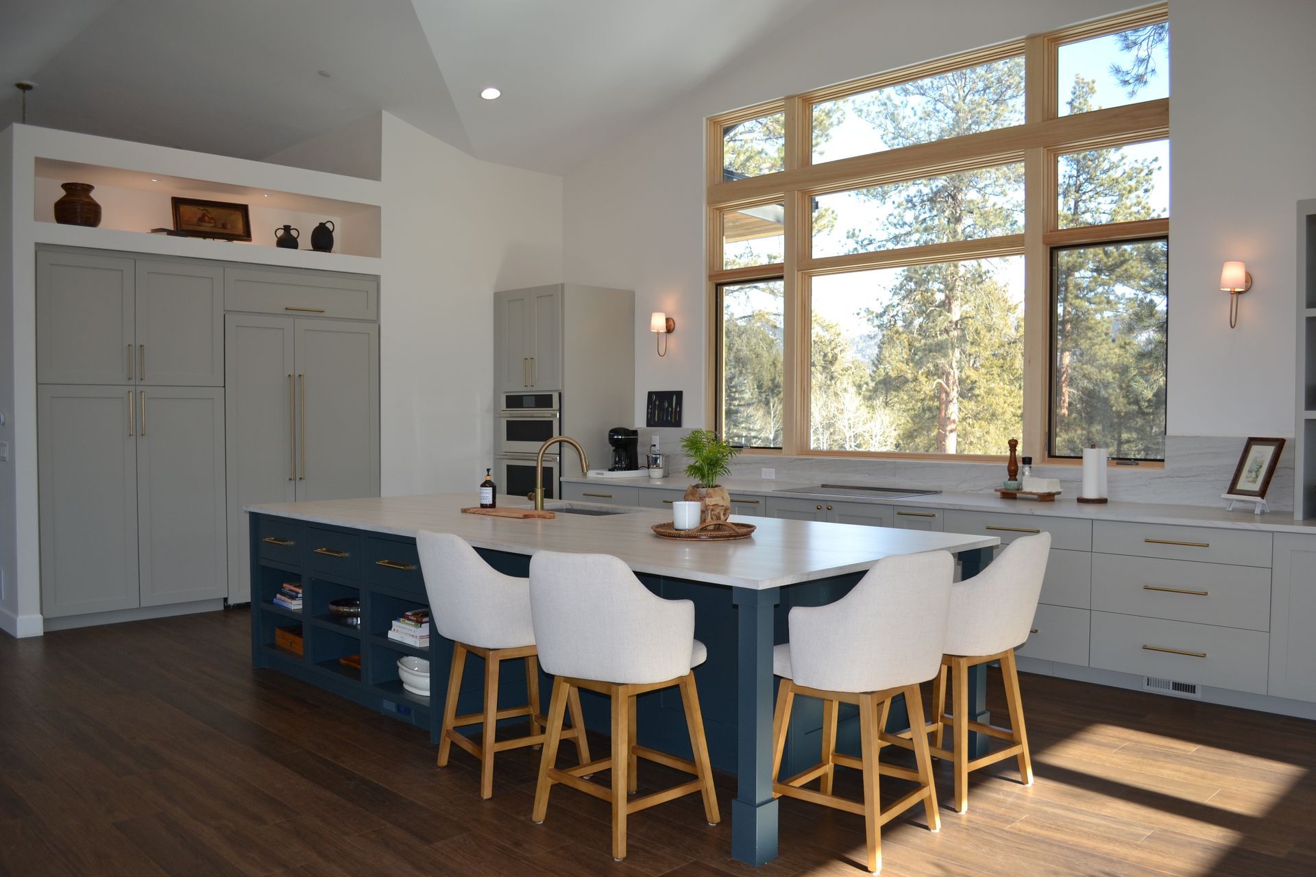 Modern kitchen with blue island, white chairs, large window with trees.