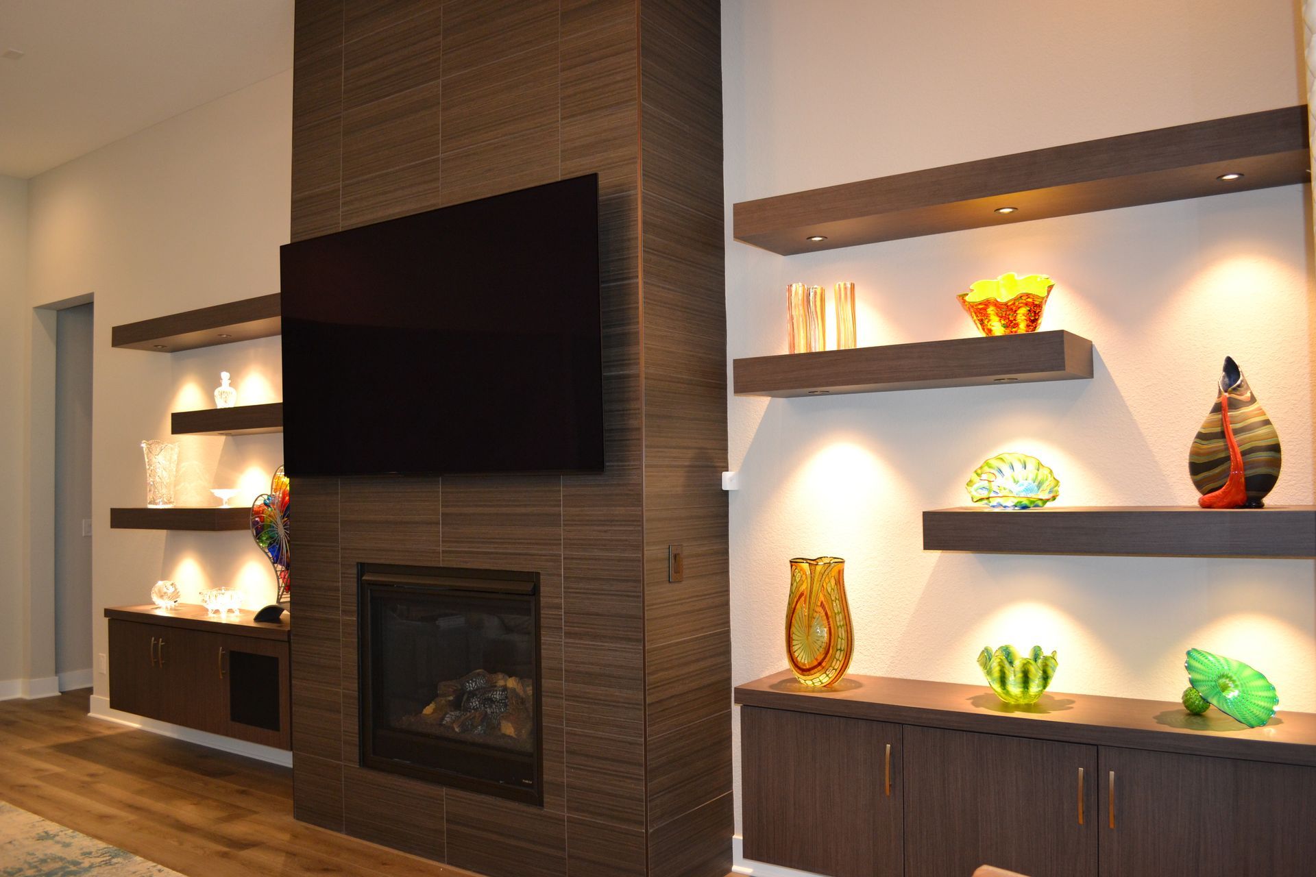 Modern living room with fireplace, TV, floating shelves with decorative items, and cabinets.