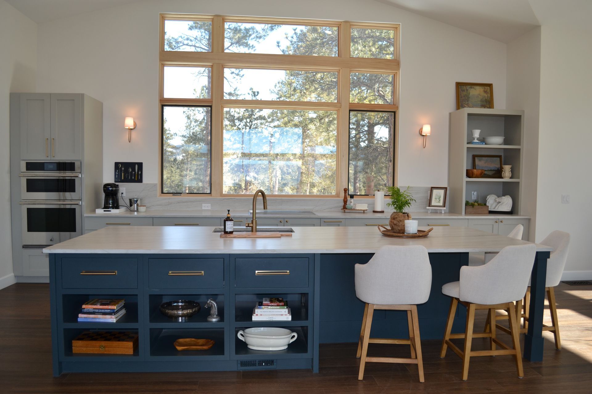 Blue kitchen island with white countertop and seating, facing a large window overlooking trees.