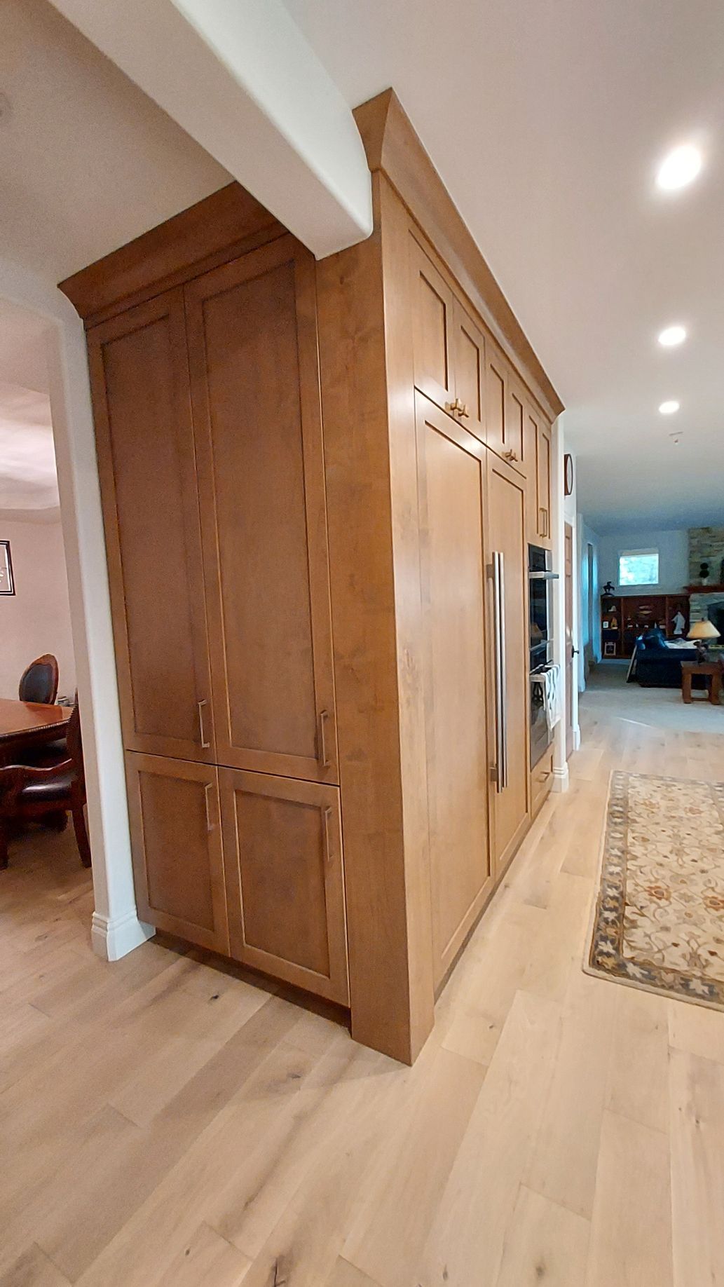 Wooden kitchen cabinetry with a built-in refrigerator and oven, separating two rooms. Light wood flooring.