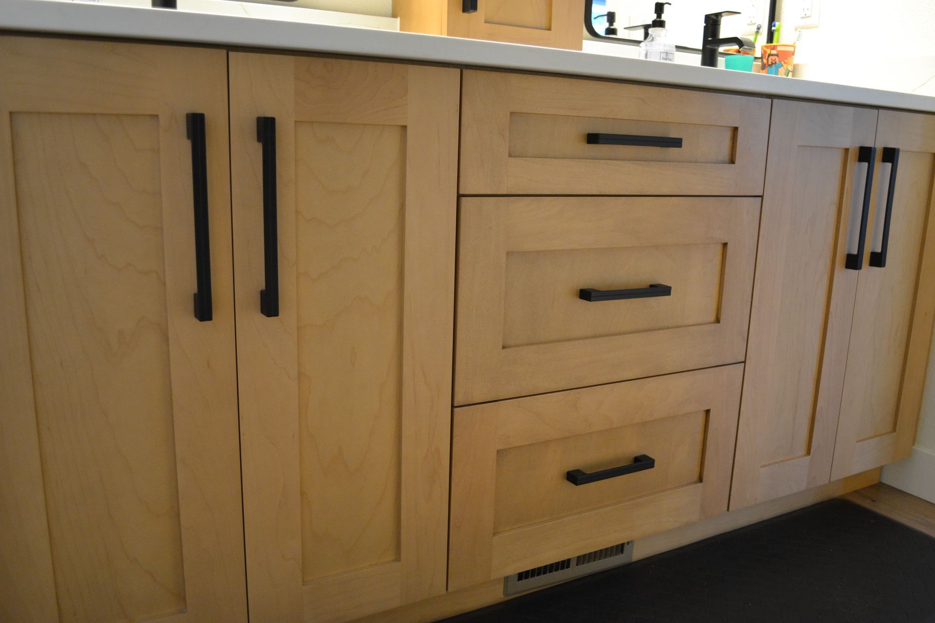 Light wood kitchen cabinets with black handles and three drawers.