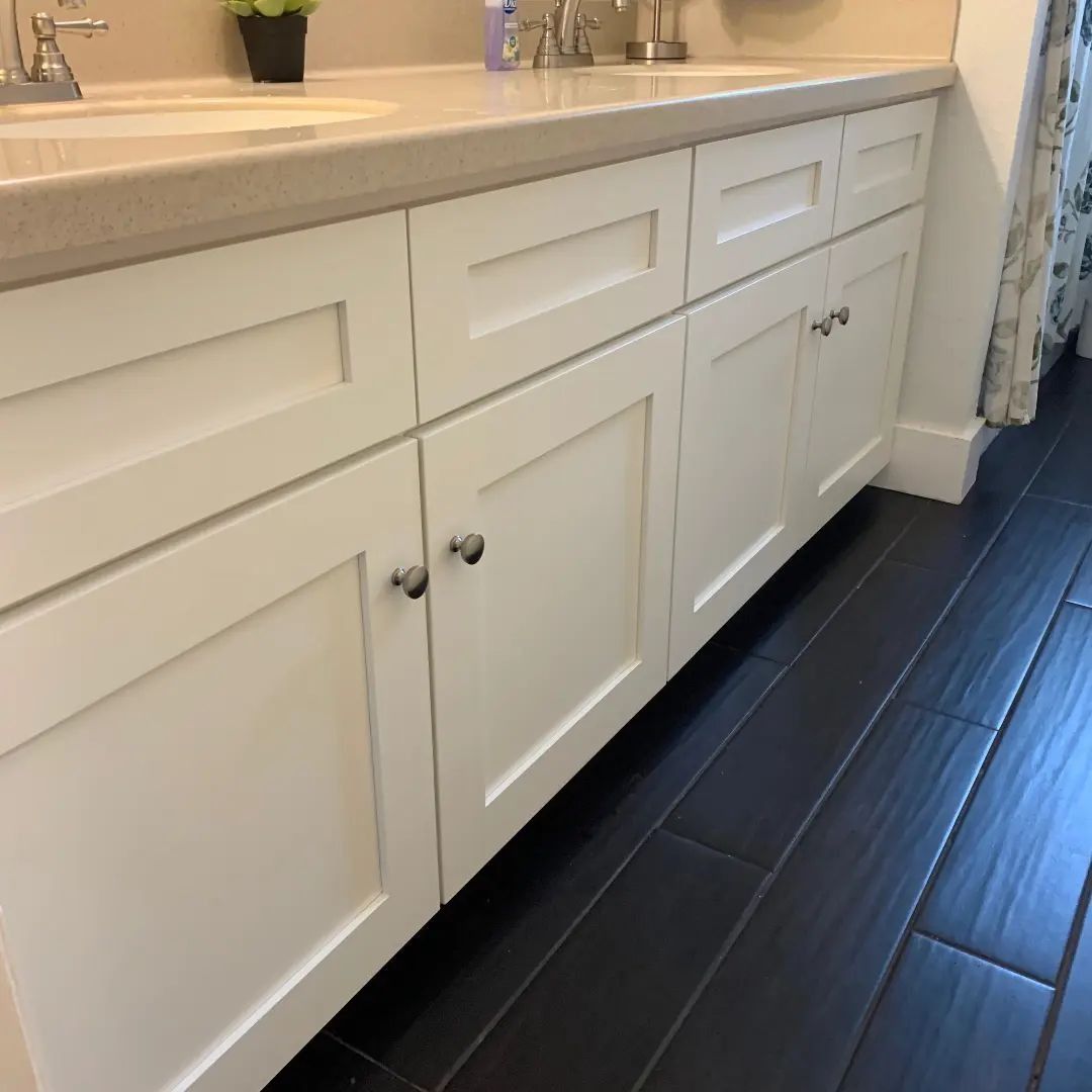 White bathroom vanity with cabinet doors and silver hardware against dark tile floor.