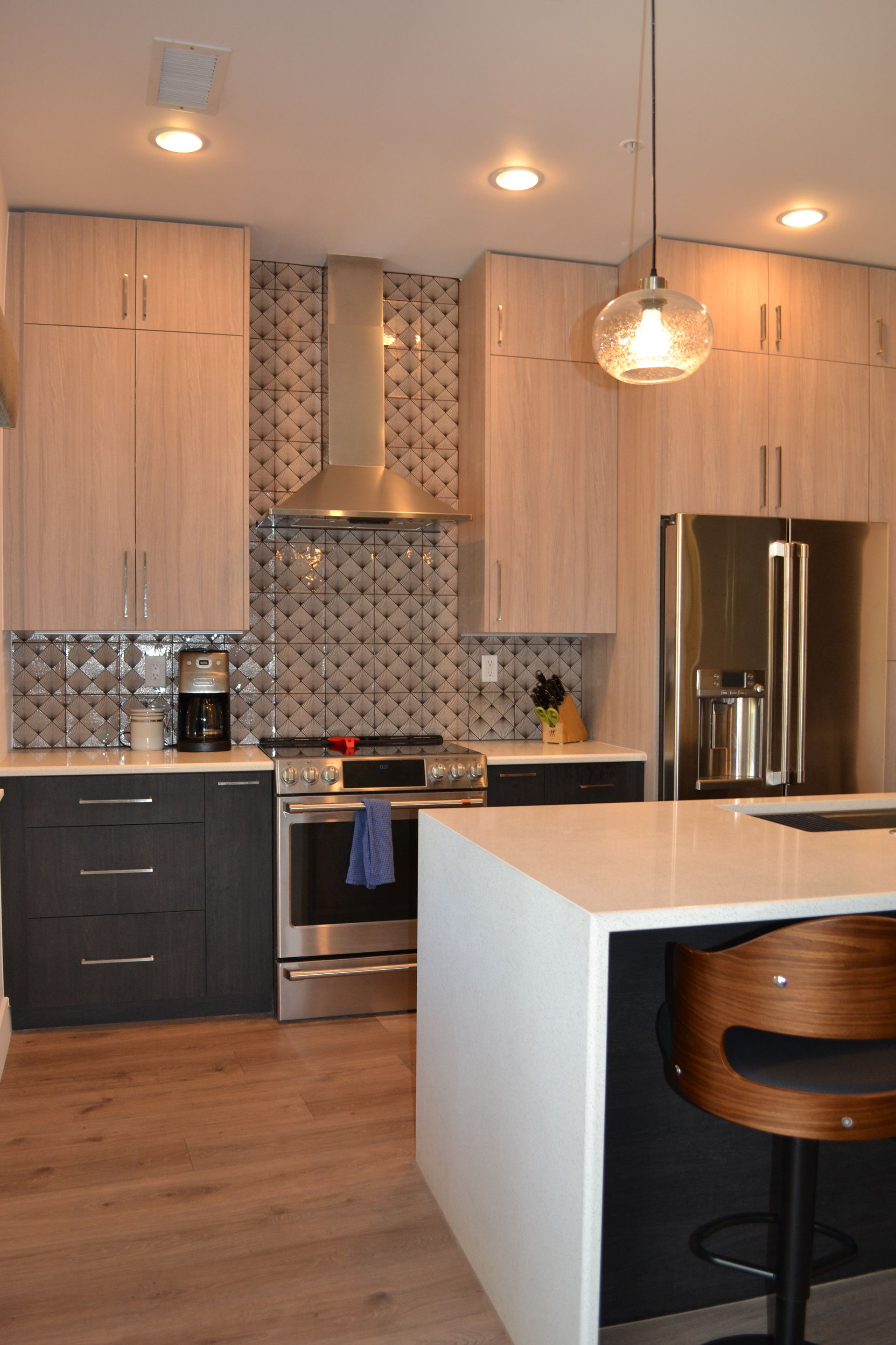 Modern kitchen with light wood upper cabinets, dark lower cabinets, stainless steel appliances, and a white countertop island.