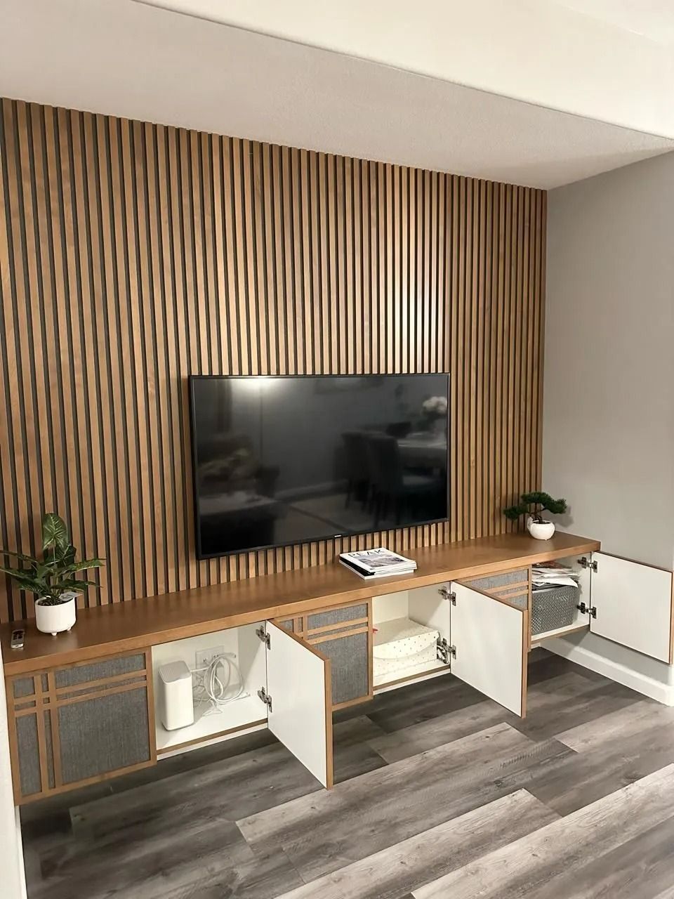 Wooden slat wall with mounted TV and low cabinet with open white doors.