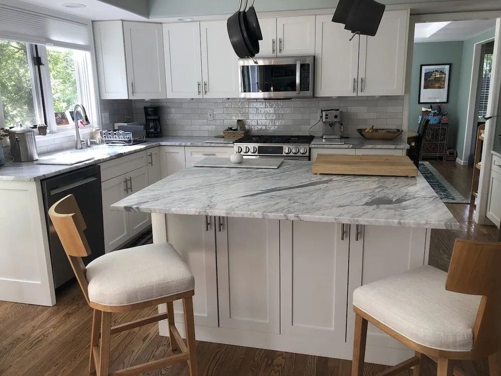 White kitchen with island, marble countertops, stainless steel appliances, and two bar stools.