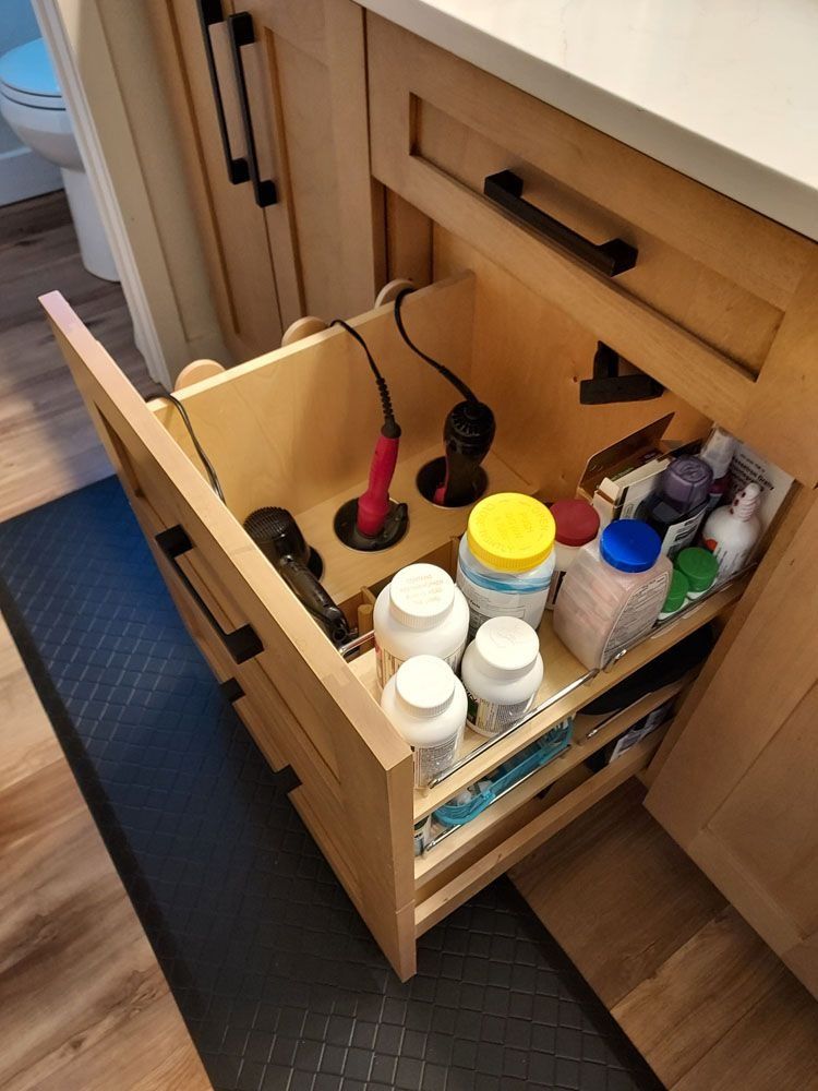 Bathroom cabinet pull-out drawer with hair tools, and toiletries. Light wood, black handles.