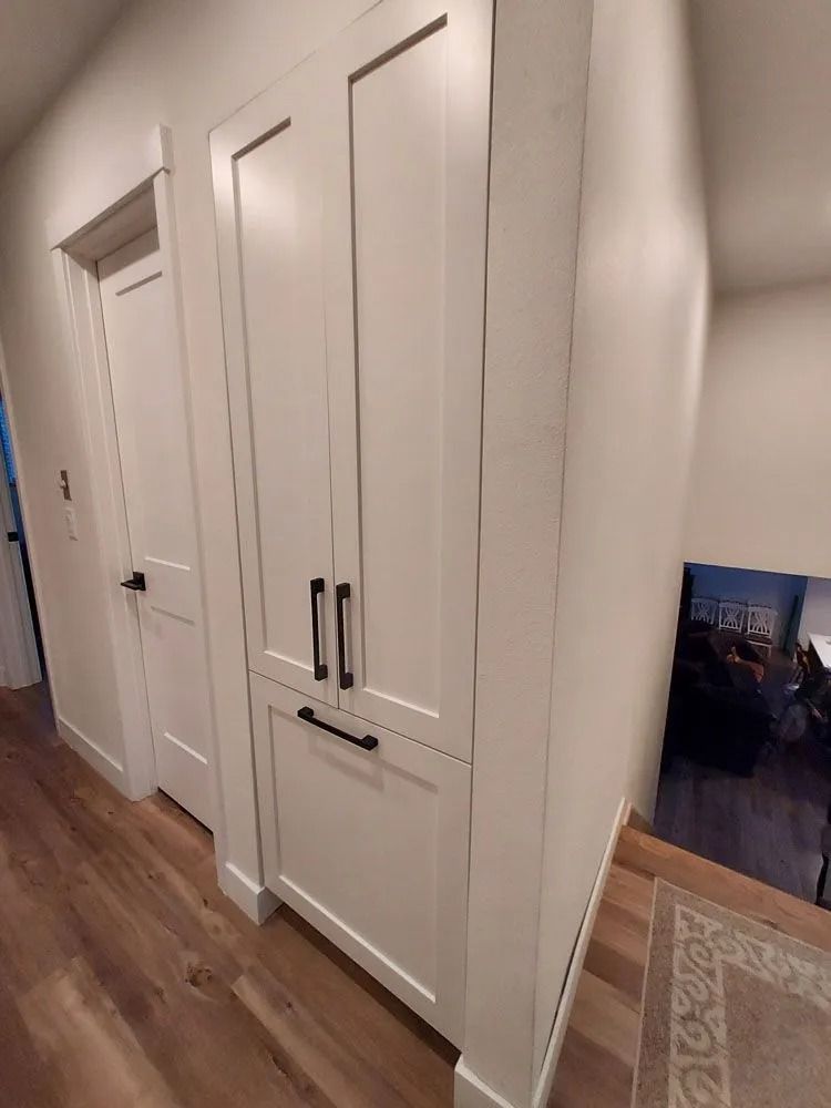 White storage cabinet with black handles next to a white door in a hallway with wood flooring.