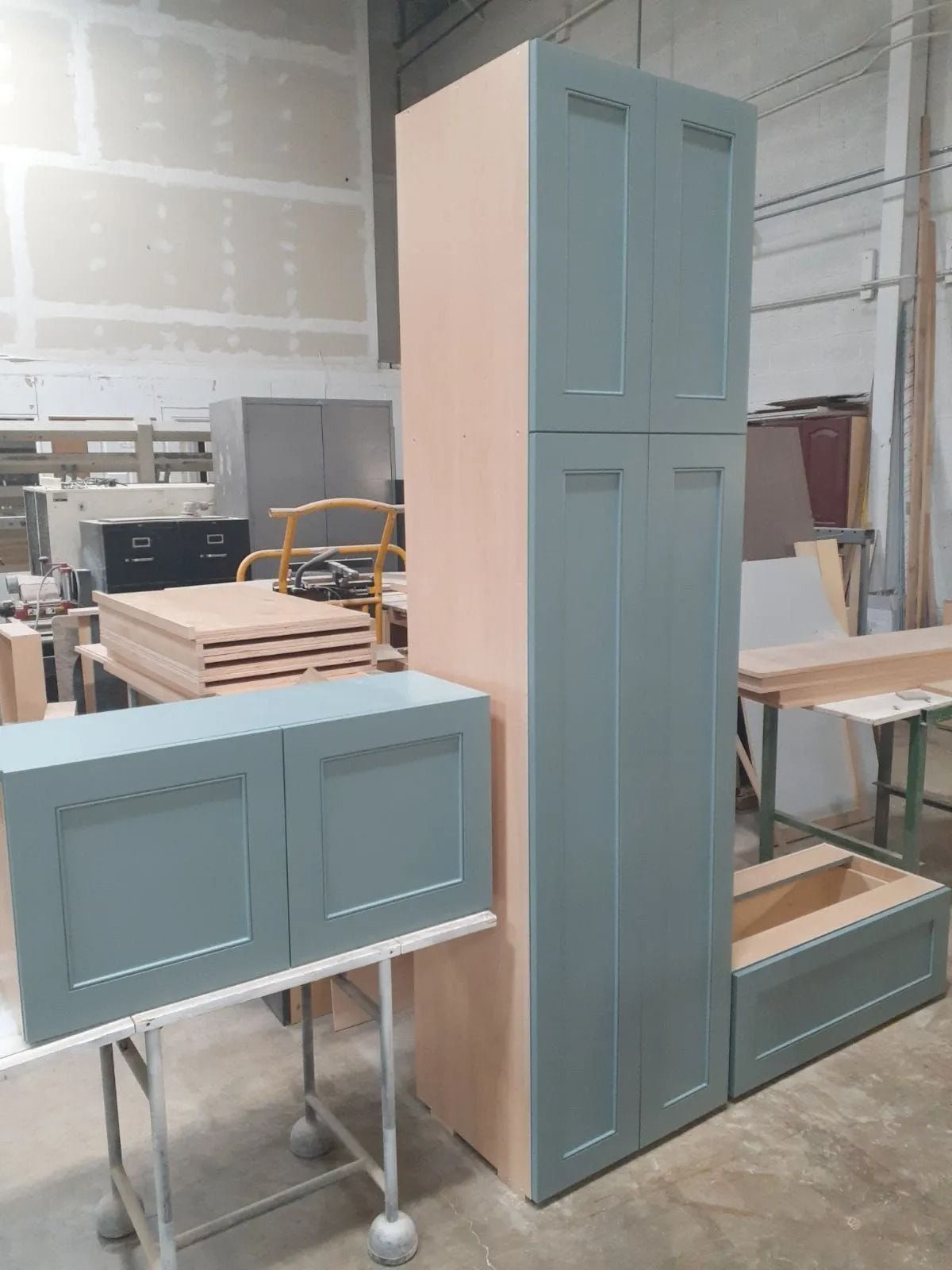 Three unfinished kitchen cabinets: two blue and one tall beige, in a workshop setting.