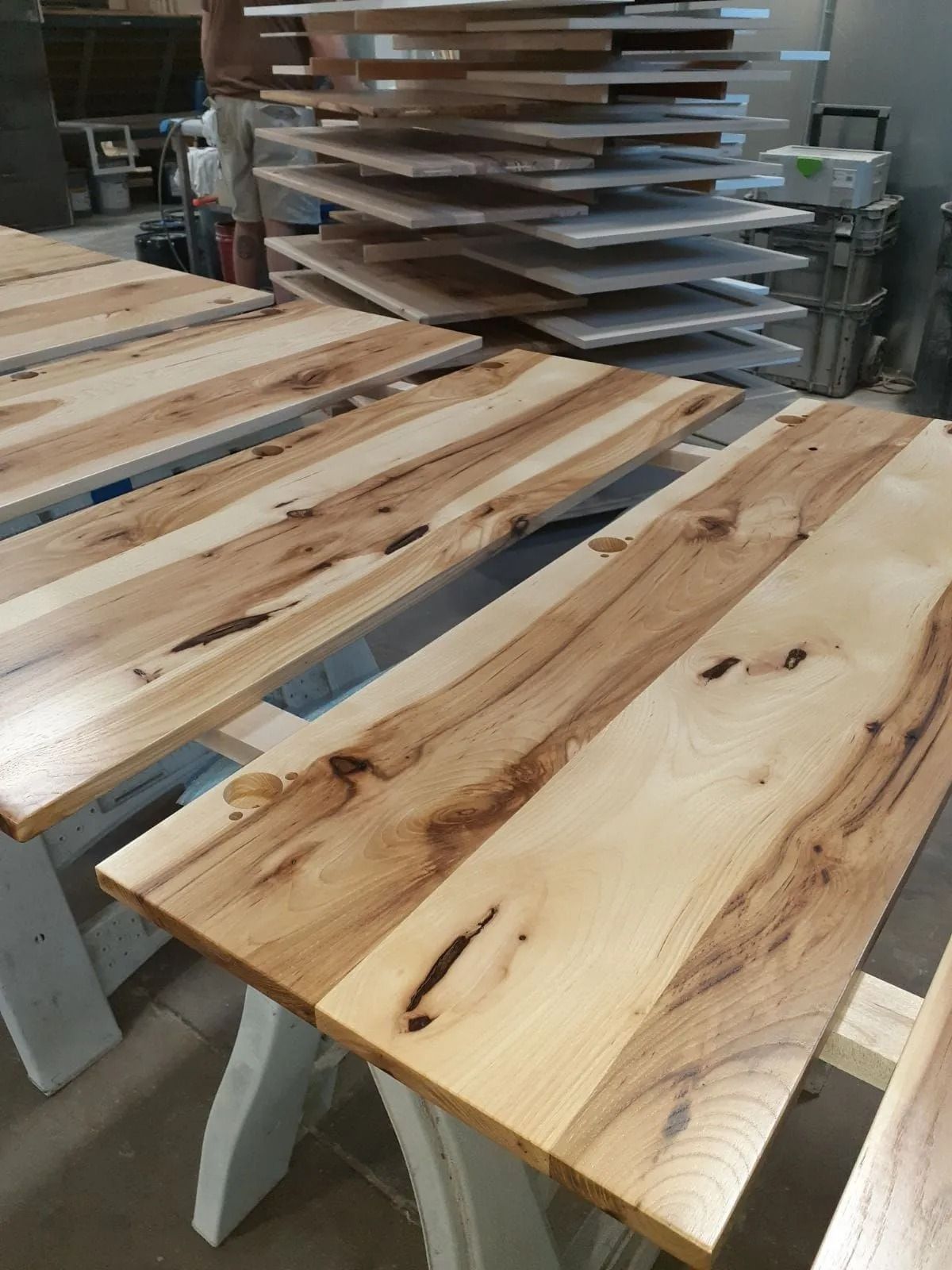 Wooden table tops in various stages of completion, with stacks in the background, set on white sawhorses.