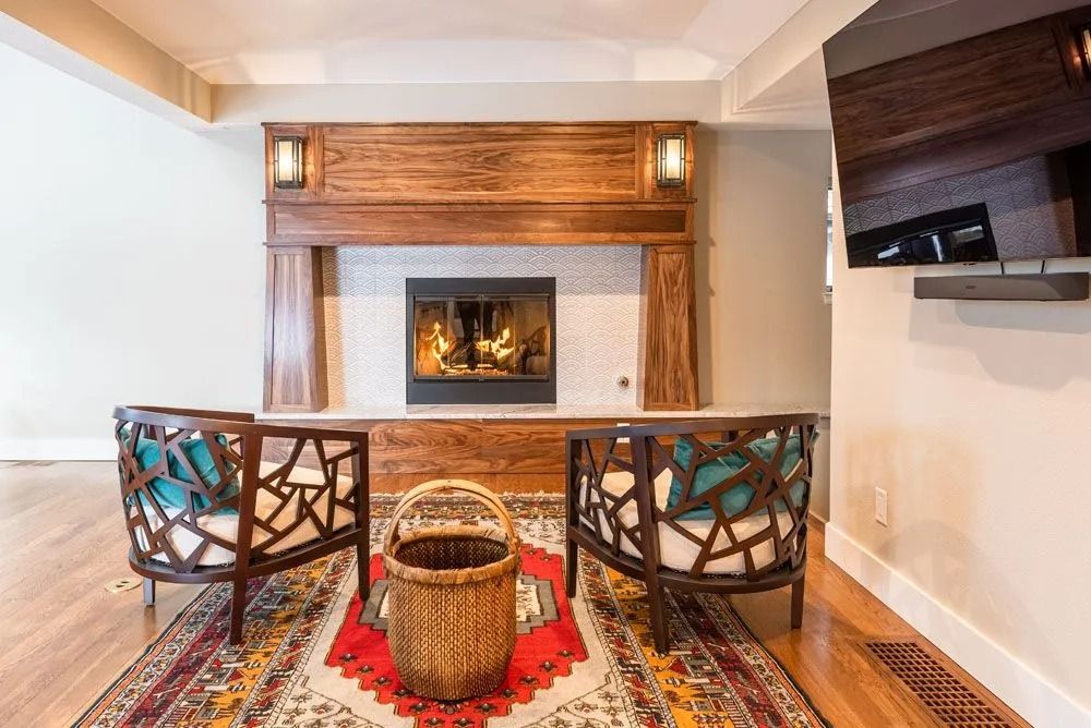 Fireplace with wooden mantel, two chairs, and a rug.