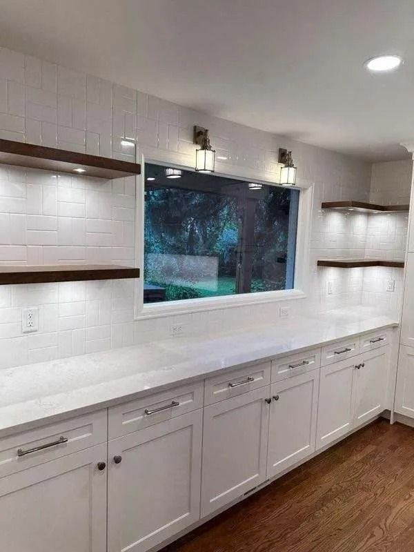 White kitchen cabinets with a long countertop, window, shelves, and lighting.