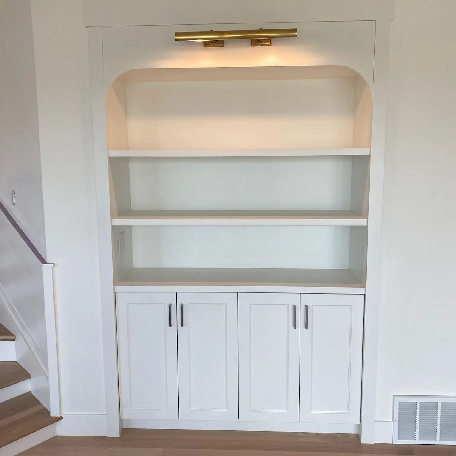 White built-in bookshelf with three shelves and lower cabinet doors. Gold light fixture illuminates the top shelf.