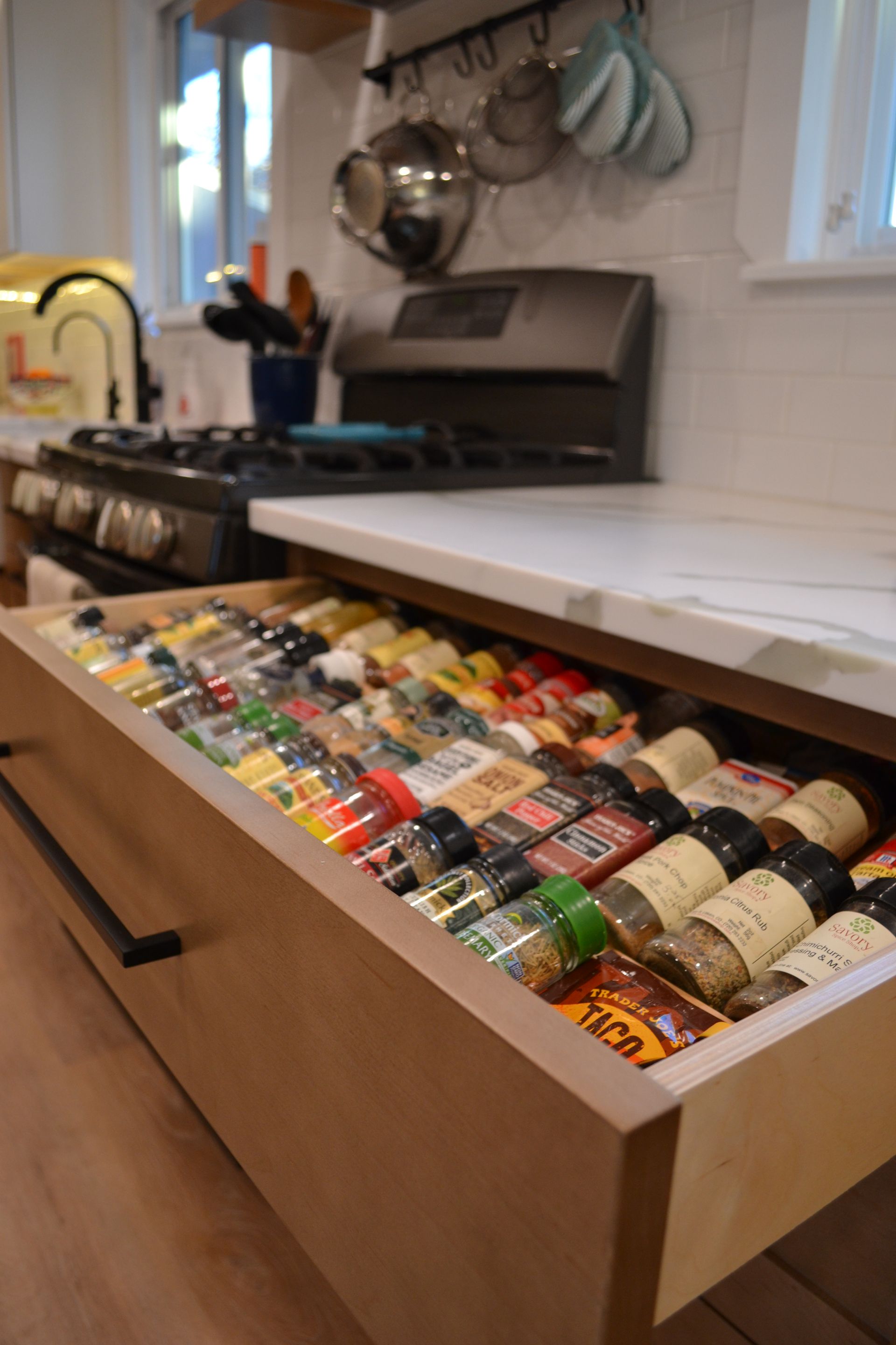 Open kitchen drawer filled with numerous spice jars, set near a stovetop and counter.