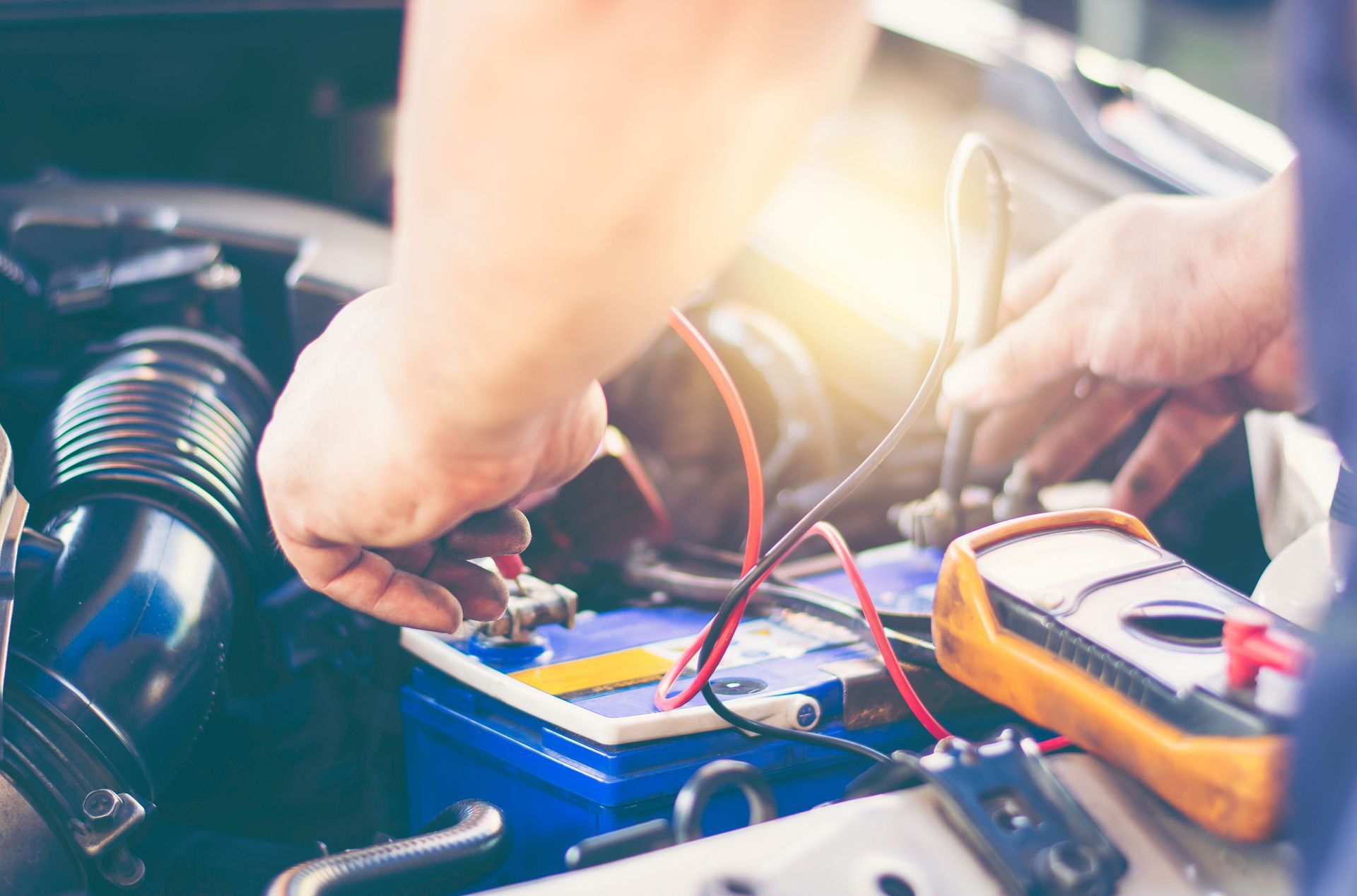 Person working on a car engine, connecting wires to a blue battery with a multimeter nearby.