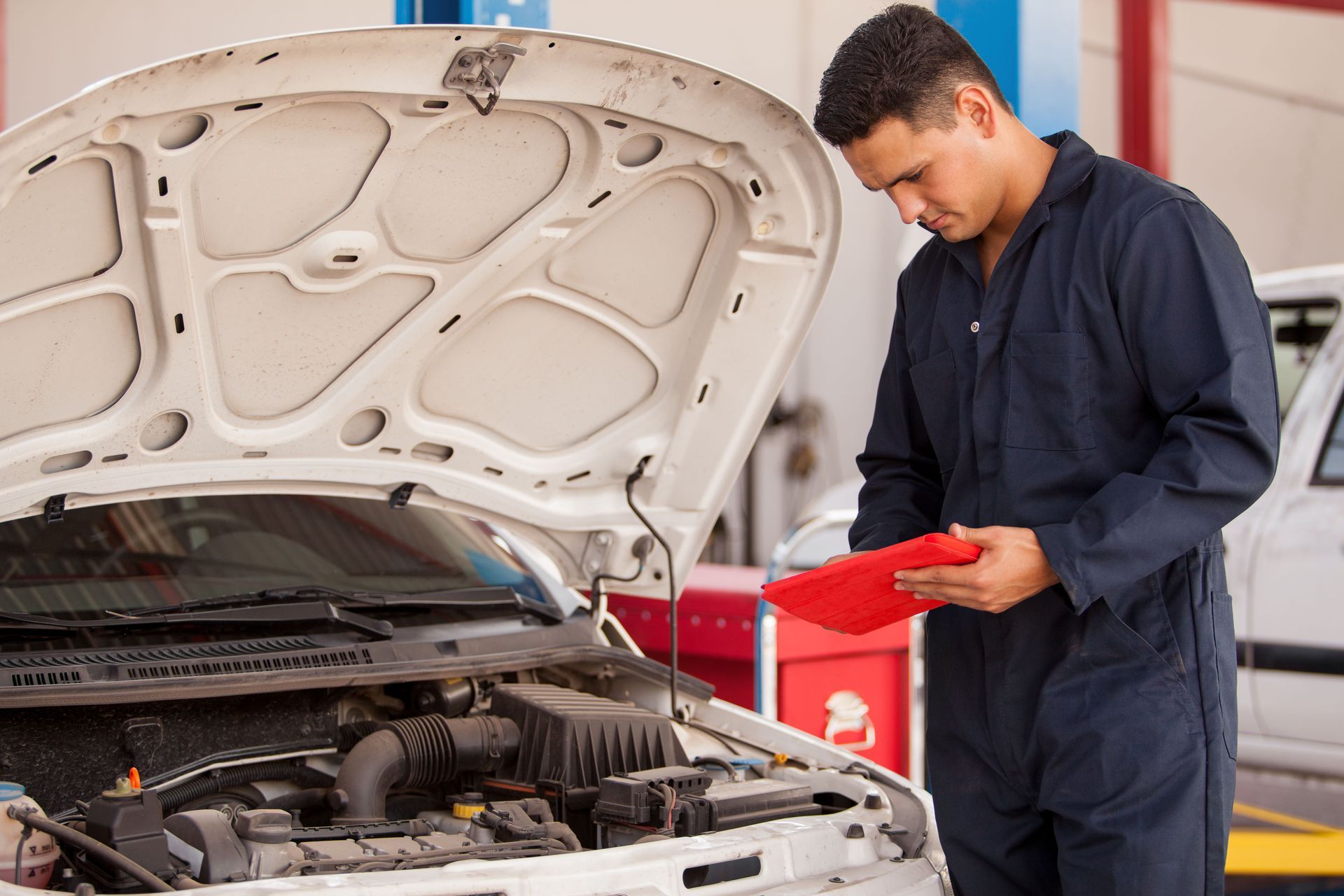 Mechanic in blue jumpsuit examining car engine, holding clipboard in a garage.