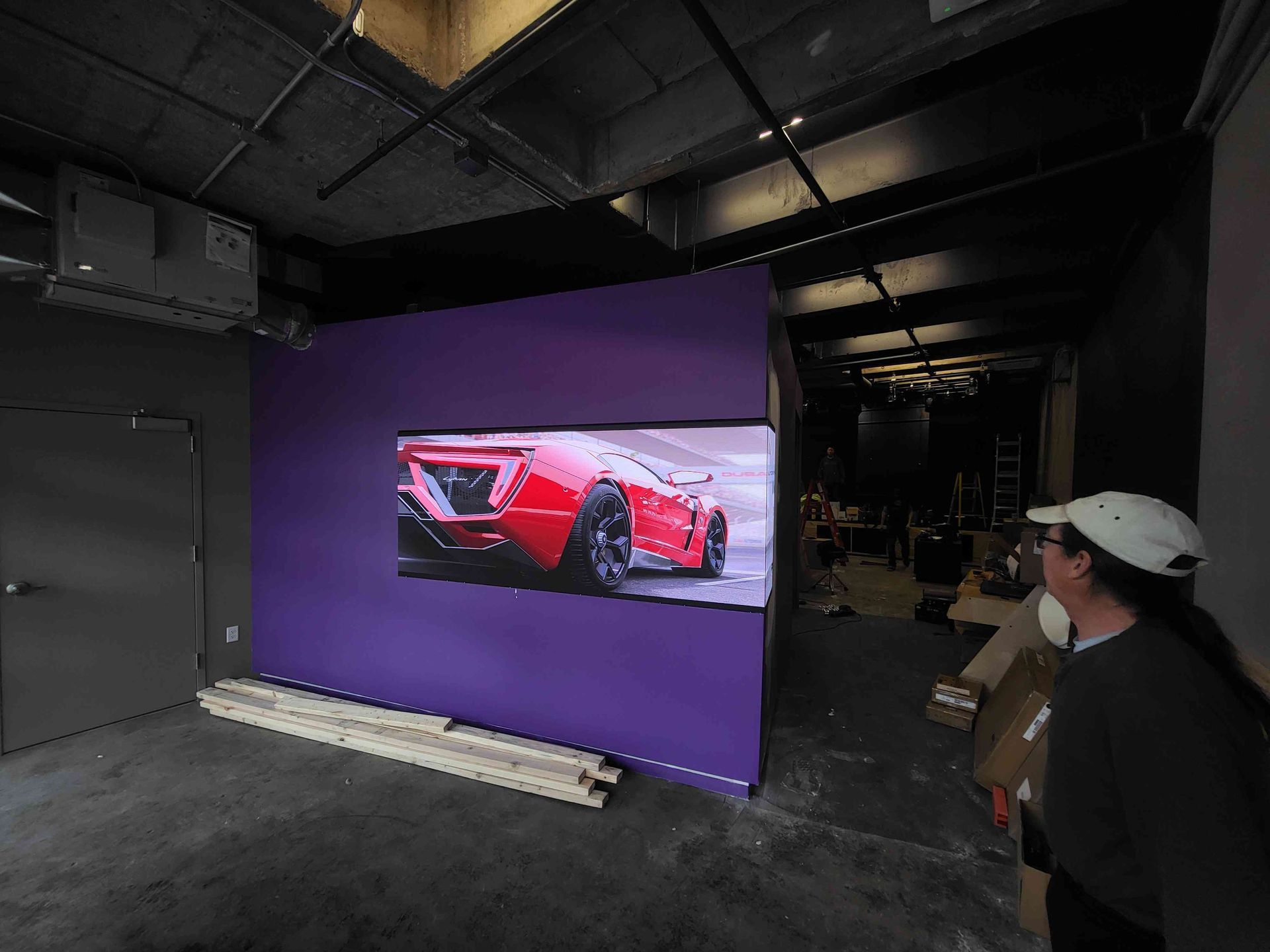 A purple wall with a car image on a screen. A person in a hat looks at it in a dark room.
