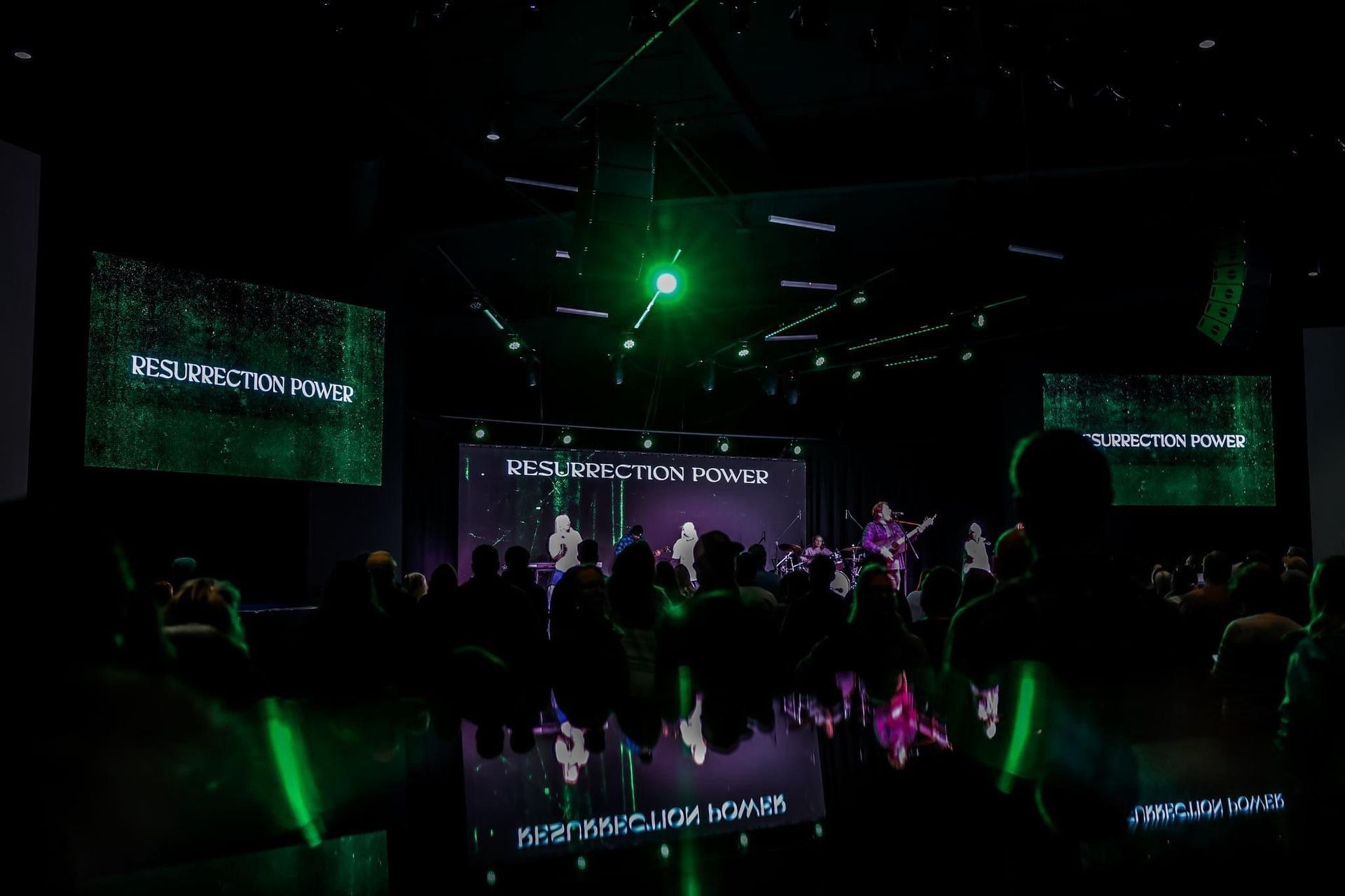 Band performing on stage with digital screens and audience in a dark venue lit with green.