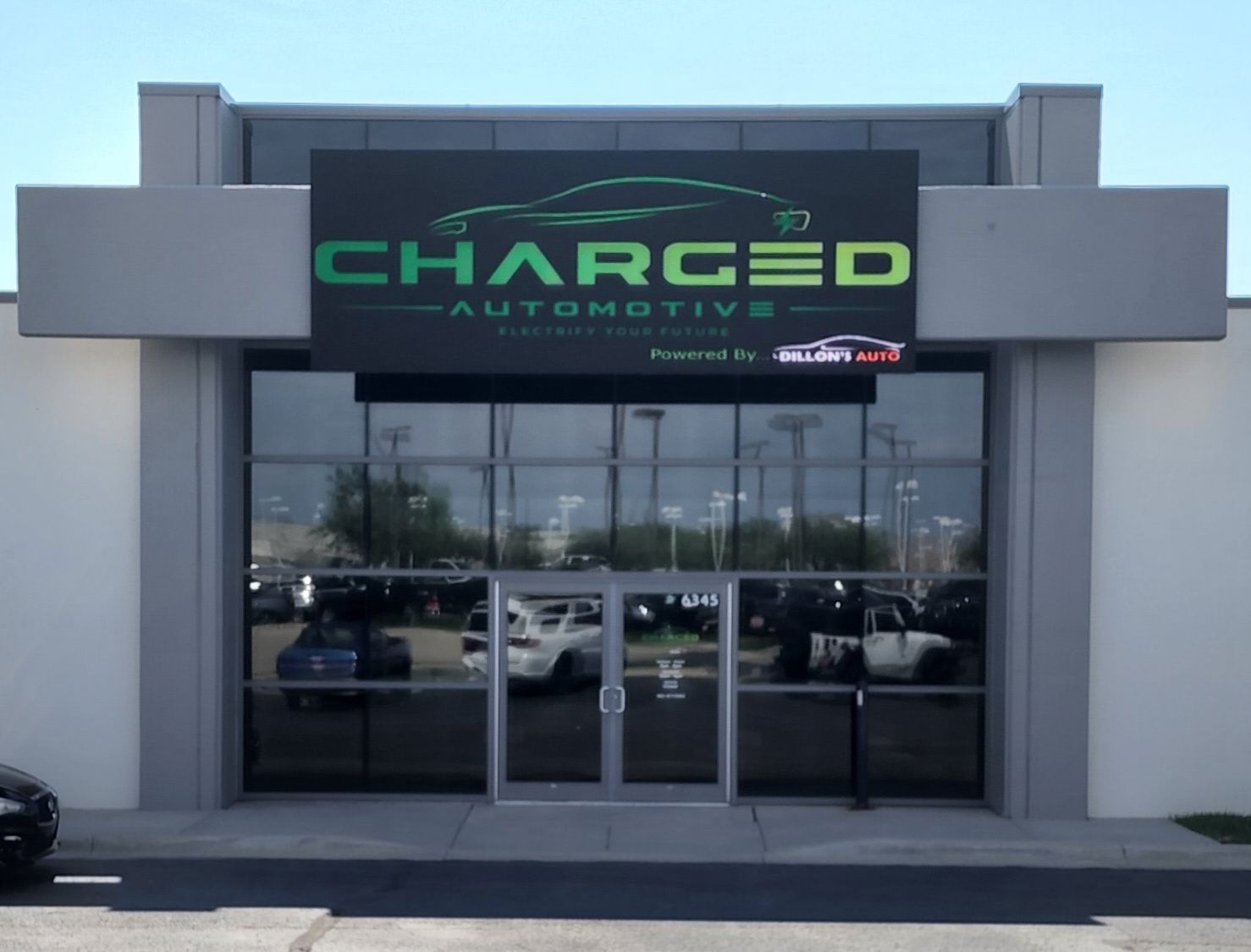 Entrance of Charged Automotive store, with green logo and black glass windows, showcasing cars inside.