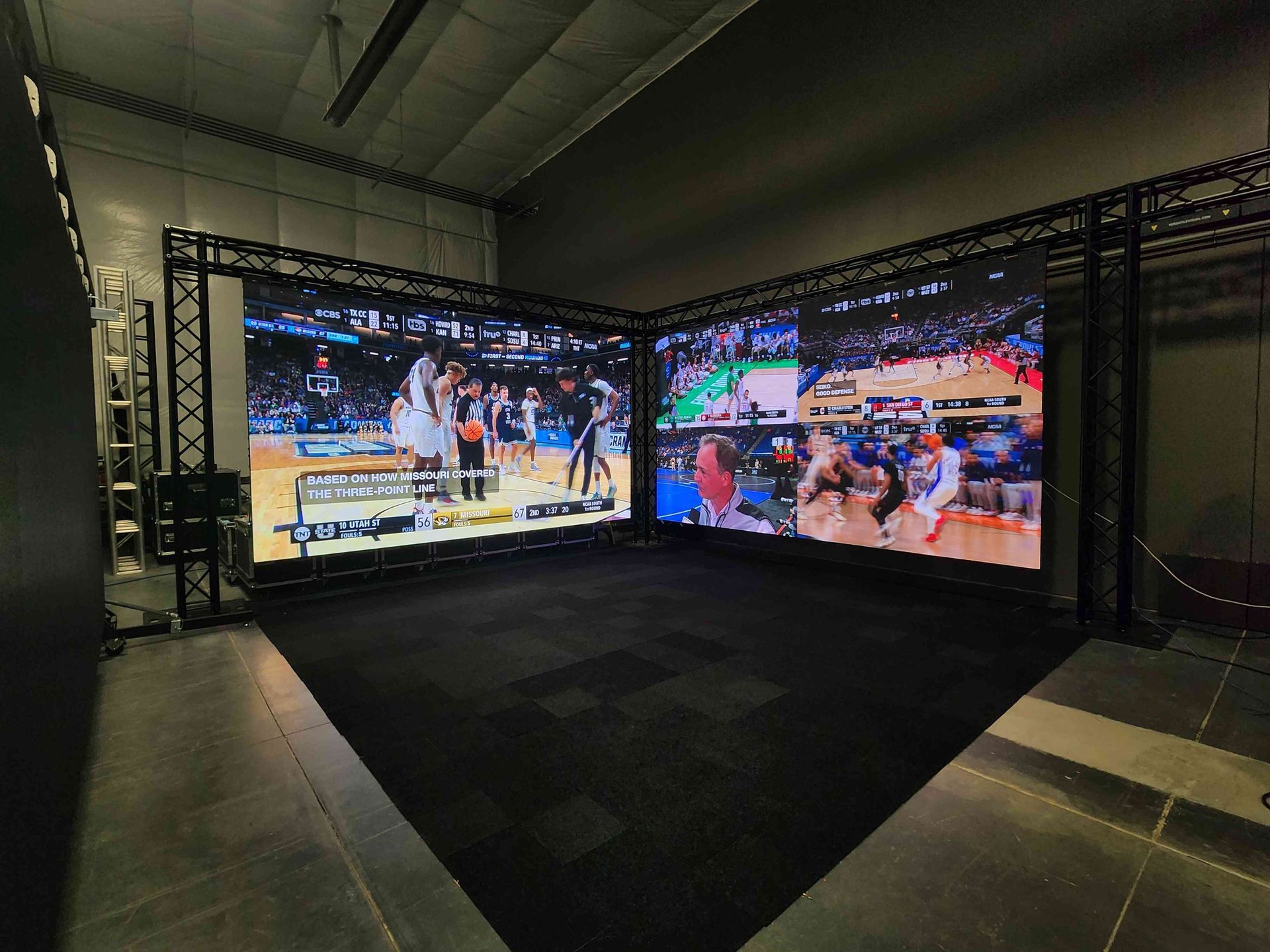 Two large screens displaying basketball game footage, set up in a corner, surrounded by black metal frames and flooring.