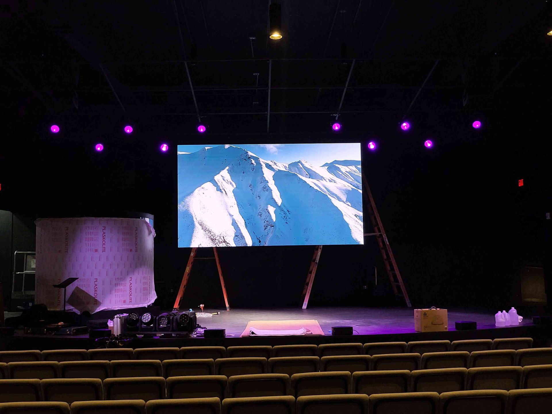 A stage with a large screen displaying mountains. Purple lights above. Empty seating in front.