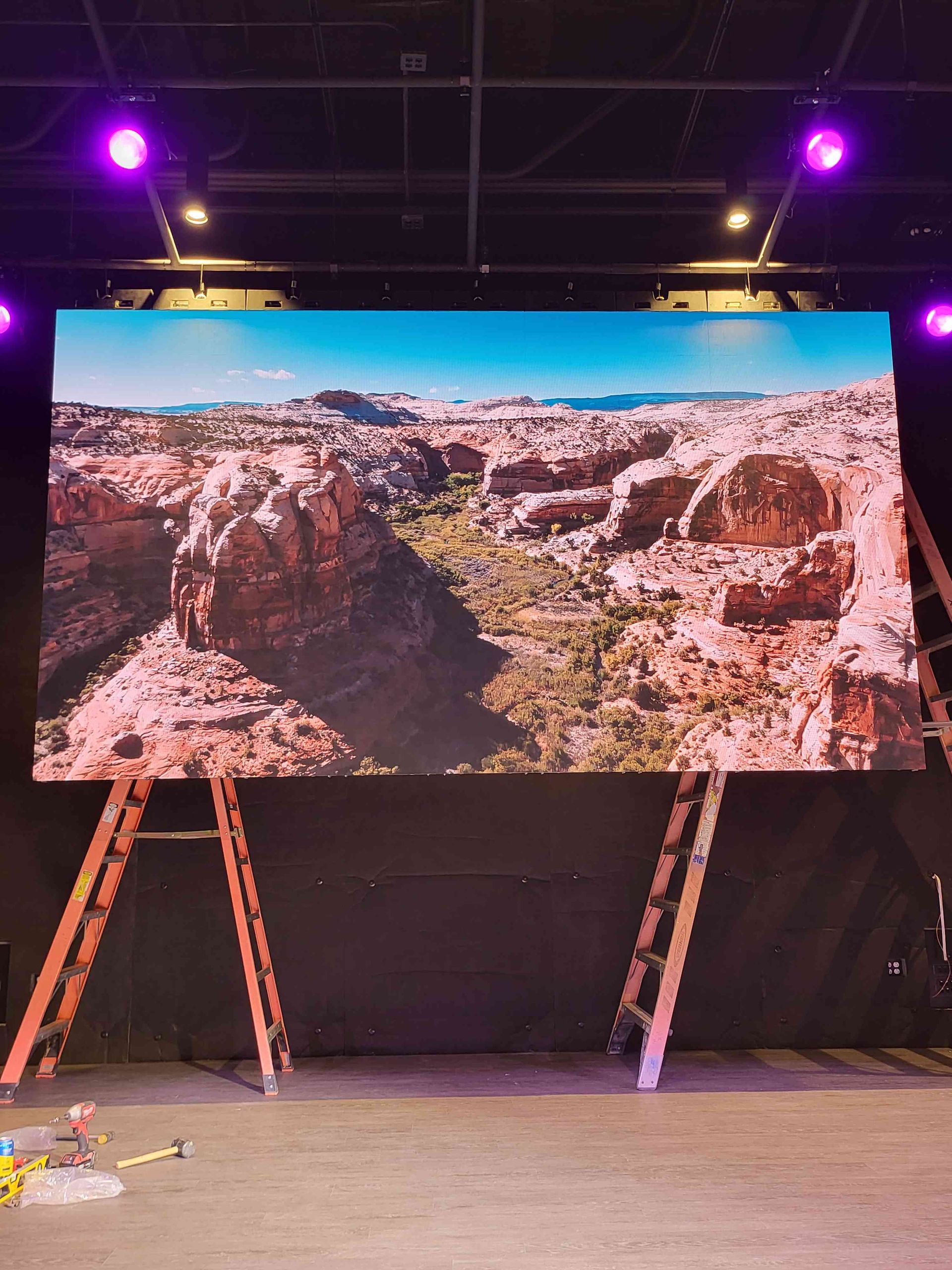 Large digital screen displaying desert landscape scene, flanked by two ladders in front of a black backdrop.