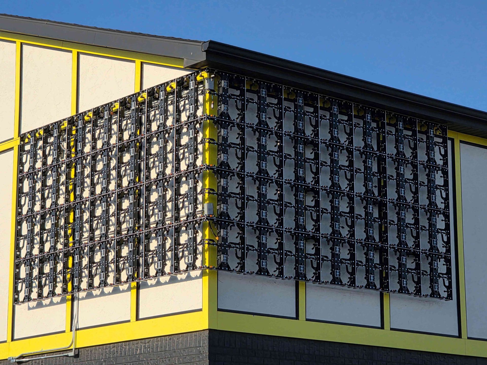 Solar panels, black grid, installed on the side of a building with yellow trim and a blue sky background.