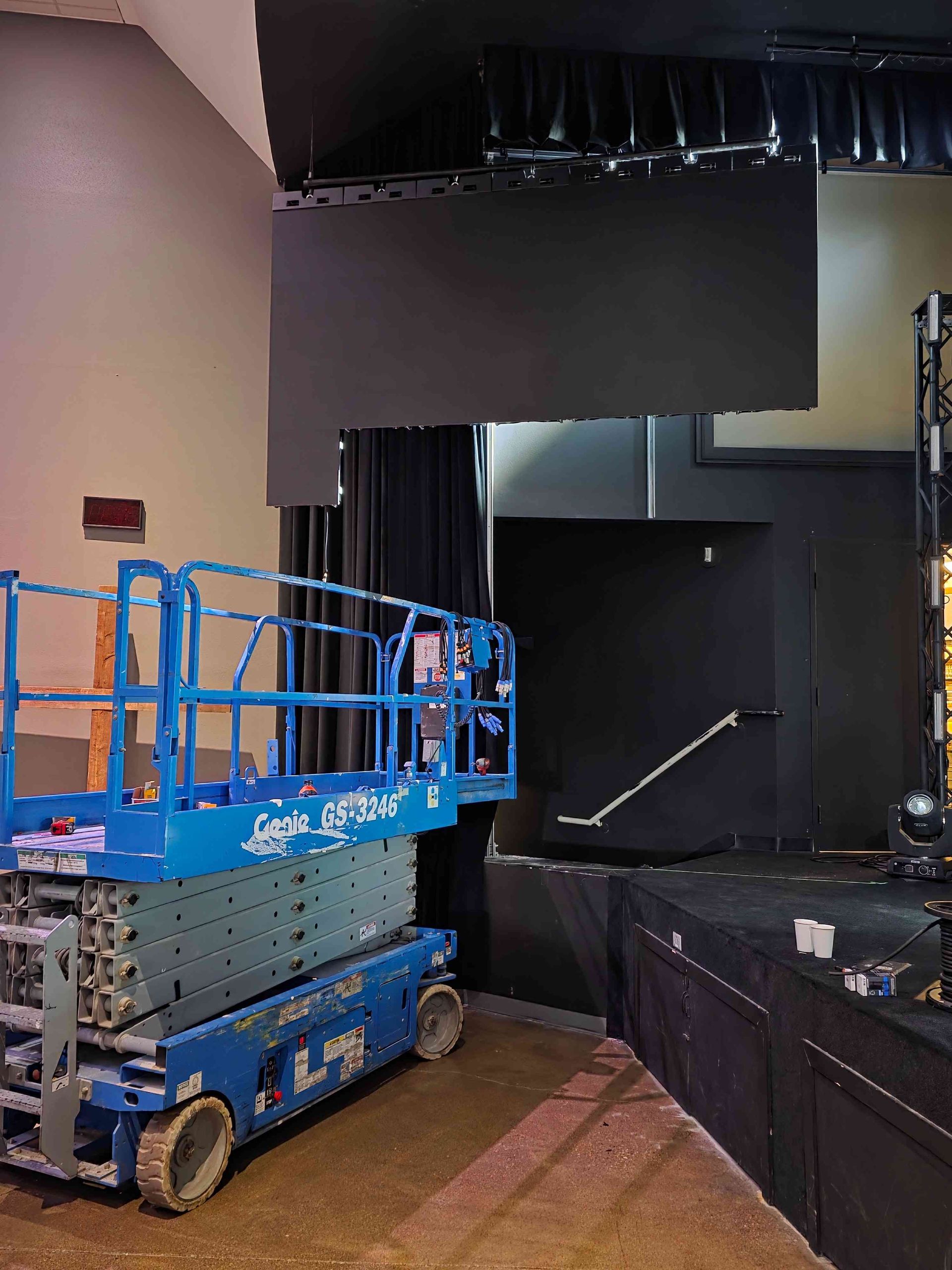 A scissor lift by a stage with a black panel above and a doorway, likely a theater during renovation.
