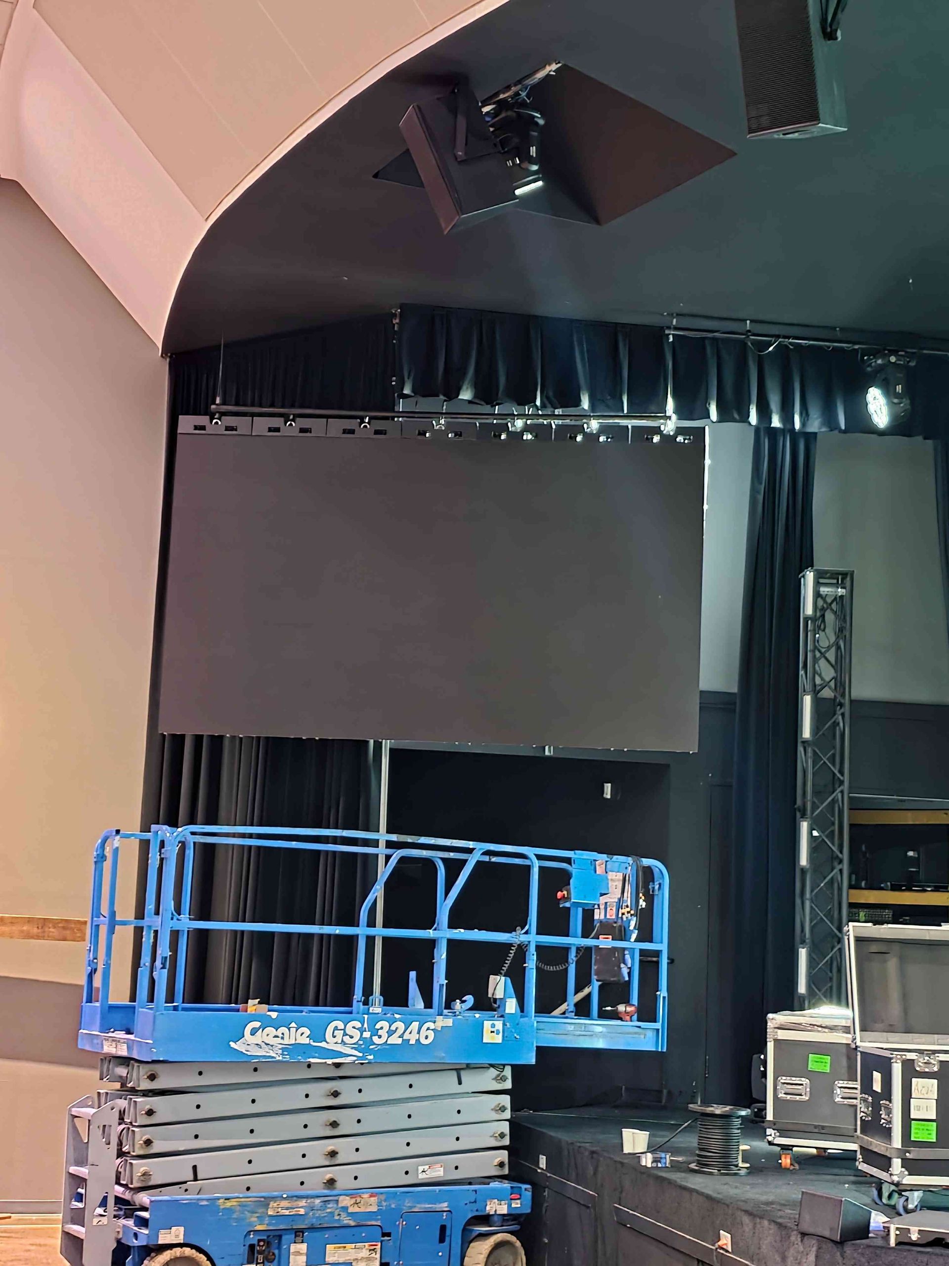 A blue lift supports a worker near a large black screen on a stage, lights above.
