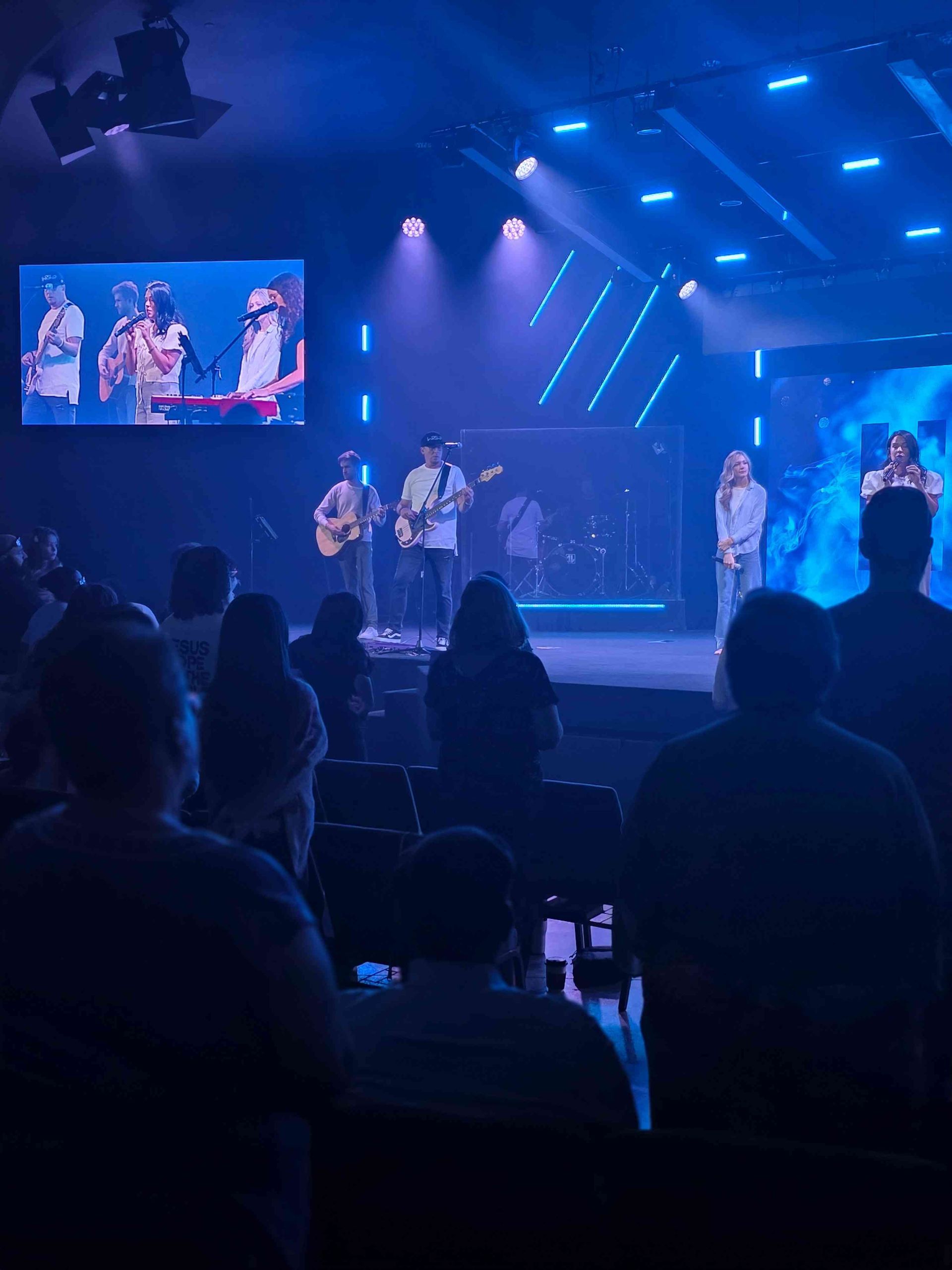 Band performing on stage under blue lights, with audience watching in a church setting.