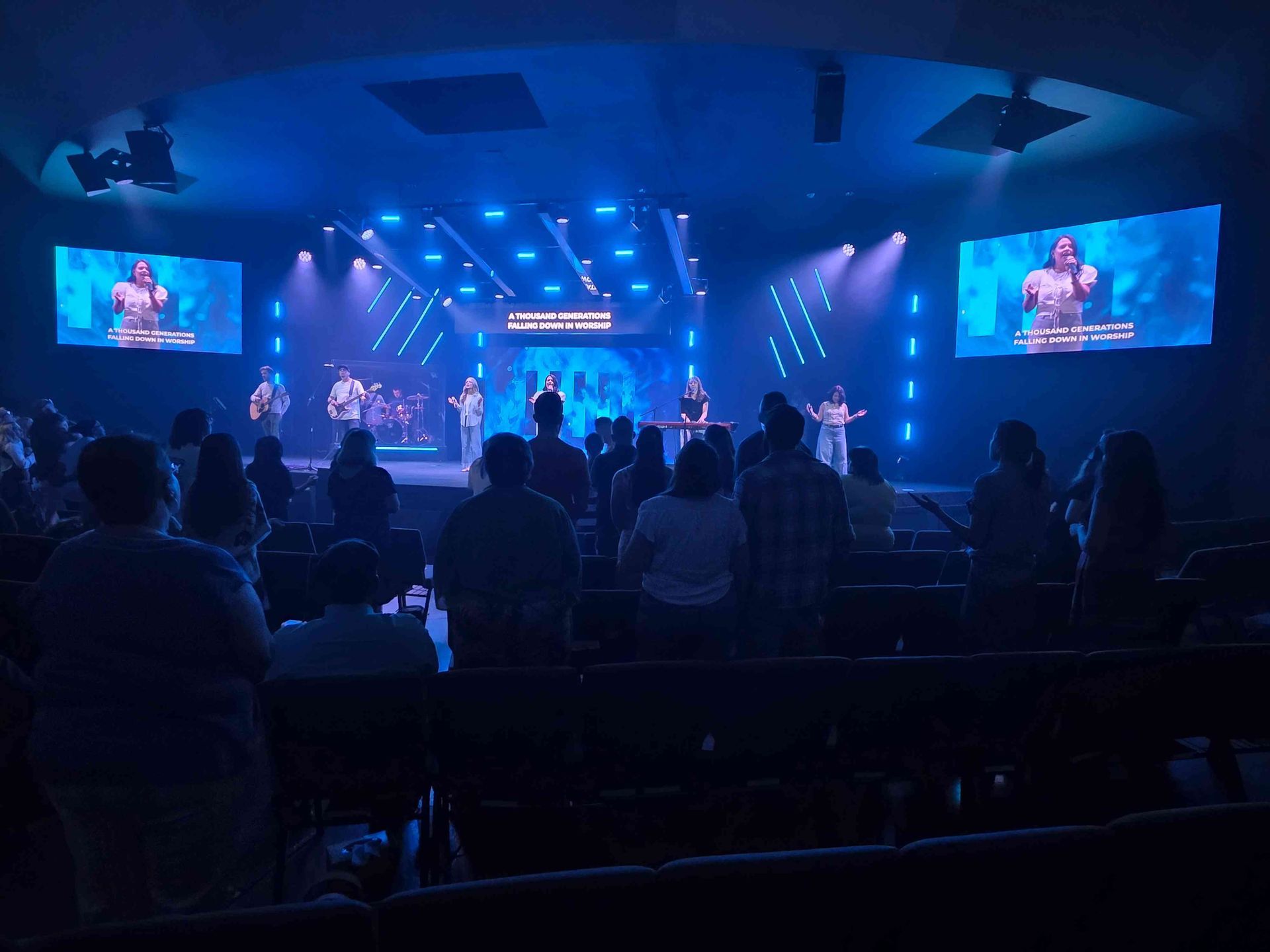 Church service with a band on stage performing for the audience. Blue lighting illuminates the stage and screens.