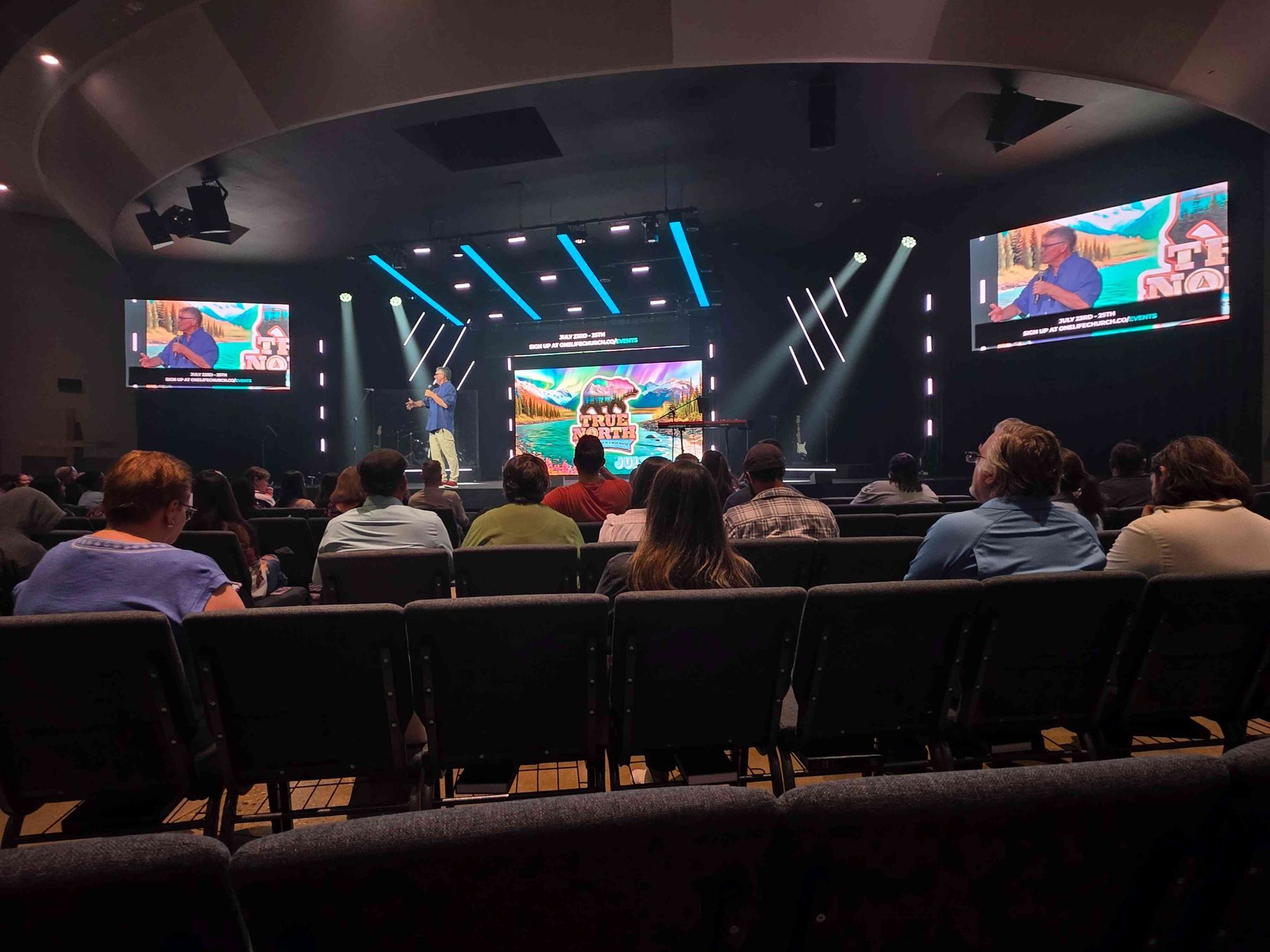 Audience at a church service. Stage with speakers, screens. Dark room, blue and white lighting. People seated.