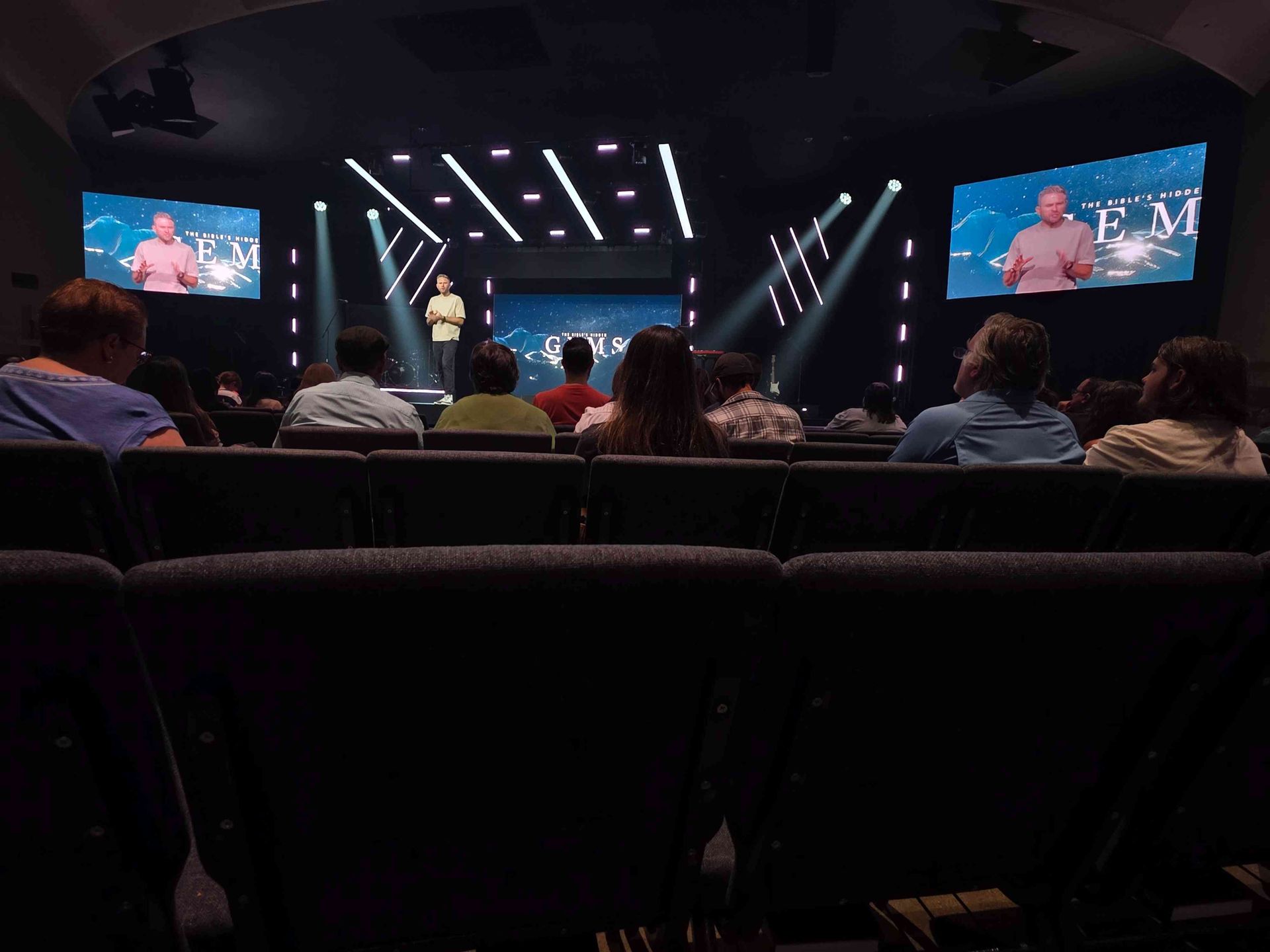 Audience seated, watching a presentation on a stage with screens, lights, and a speaker in a large venue.