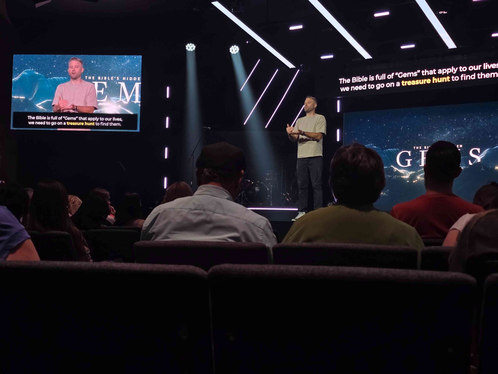 A speaker on stage, addressing an audience in a church. A screen displays text and a man speaking.