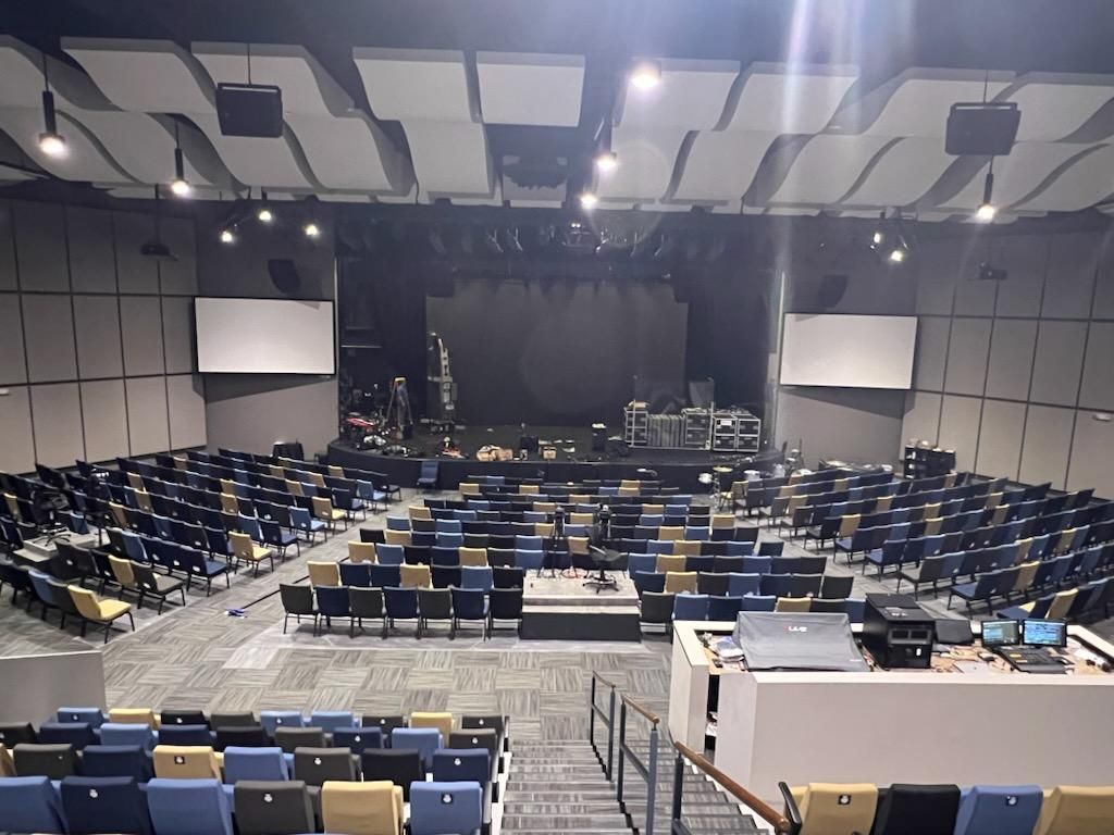 Auditorium with rows of blue and beige seats facing a stage. Sound equipment is set up on stage.