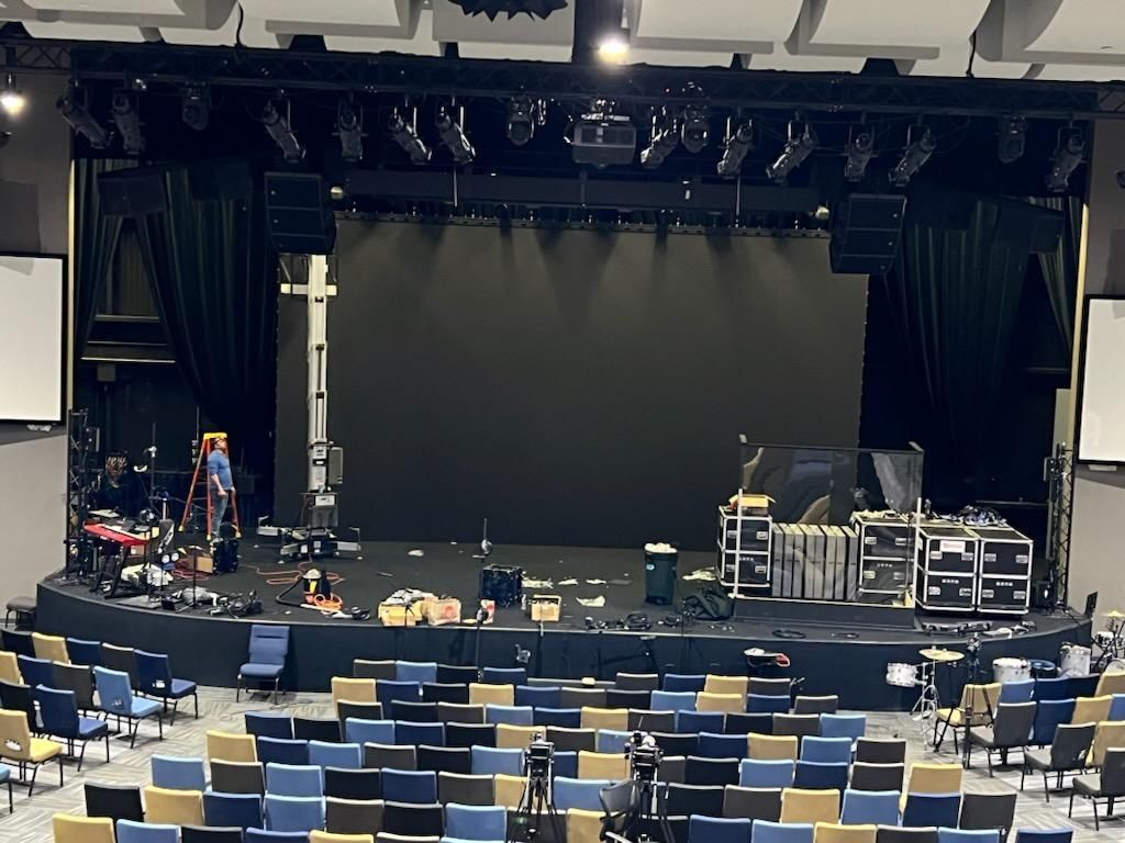 A theater stage with black curtains, lights, and equipment, facing an audience of blue and tan seats.