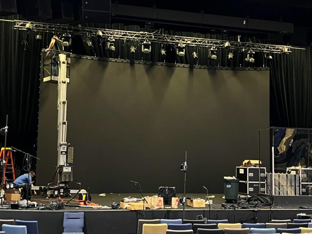 Stage setup with large black screen, lighting rig, equipment, and workers in an empty auditorium.