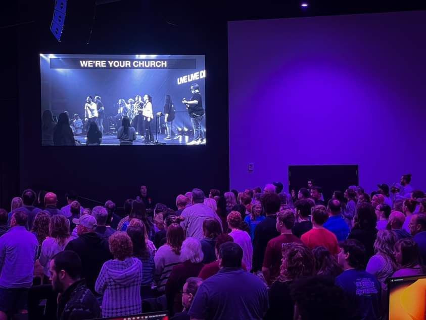 Audience in a large room watching a video on a screen. Text reads 