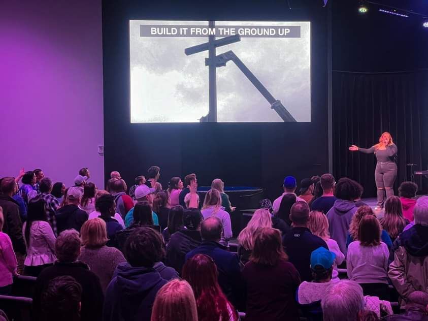 Audience watches a speaker on stage in a theater; screen shows 