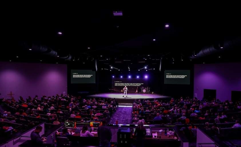 A person speaking on stage in a large auditorium with a purple hue; audience seated.
