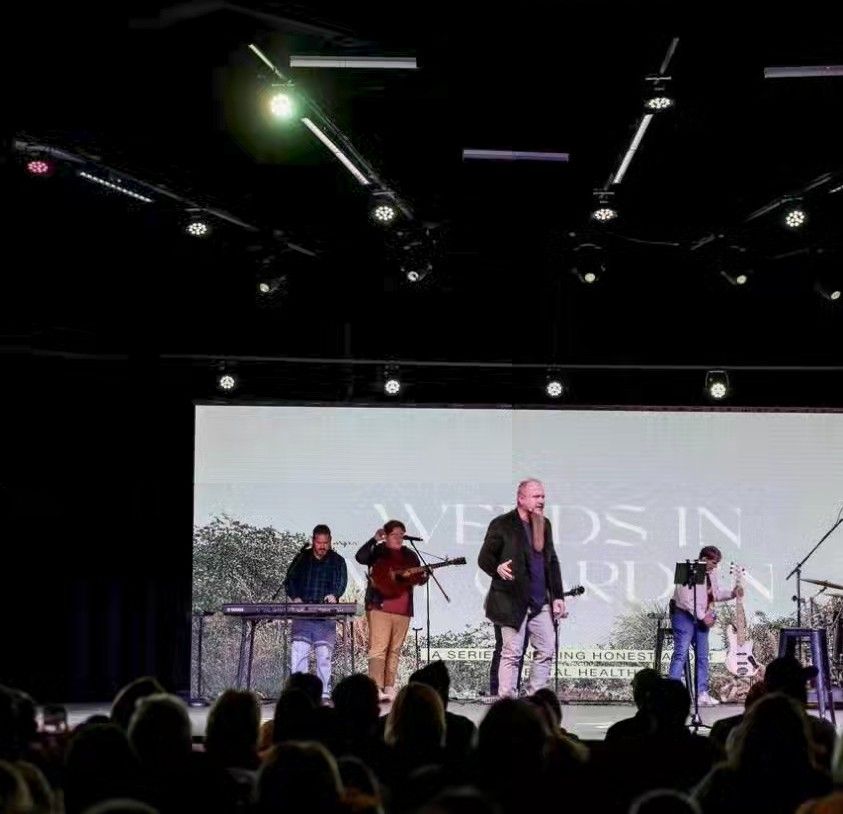 A band performs on stage with a large screen backdrop; audience in foreground.