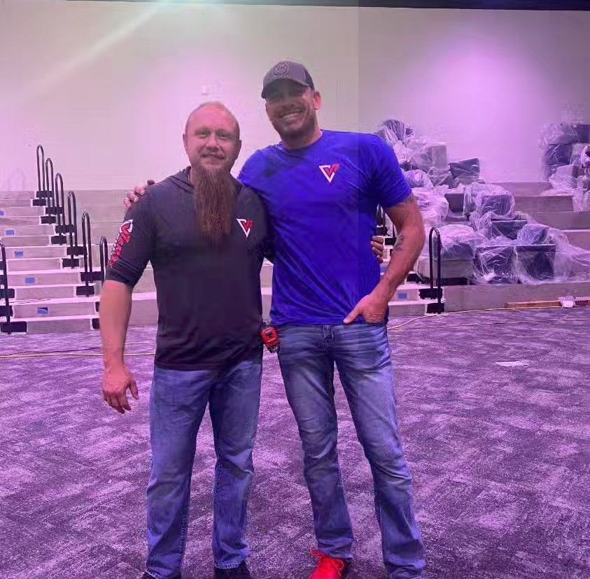 Two men stand posing, one with a beard, the other in a blue shirt. Inside a venue.