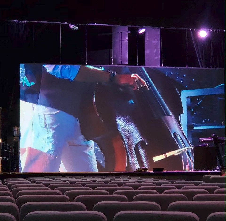Stage with a large screen displaying a musician playing a cello. Rows of empty seats in the foreground.