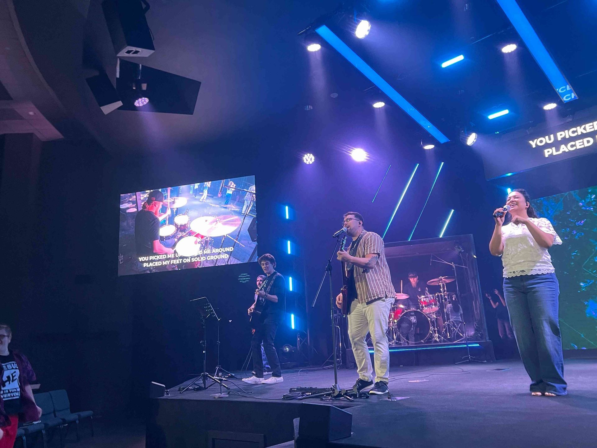 A band performs on a stage, with a singer, guitarist, bassist, and vocalist. Blue lighting and a screen display lyrics.