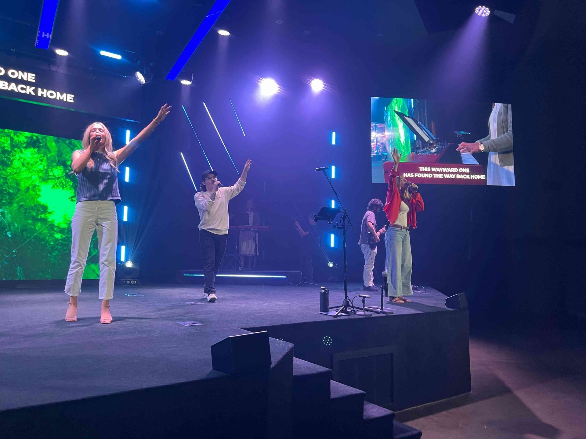 A band performs on stage under blue and white lights. Three singers raise their hands.